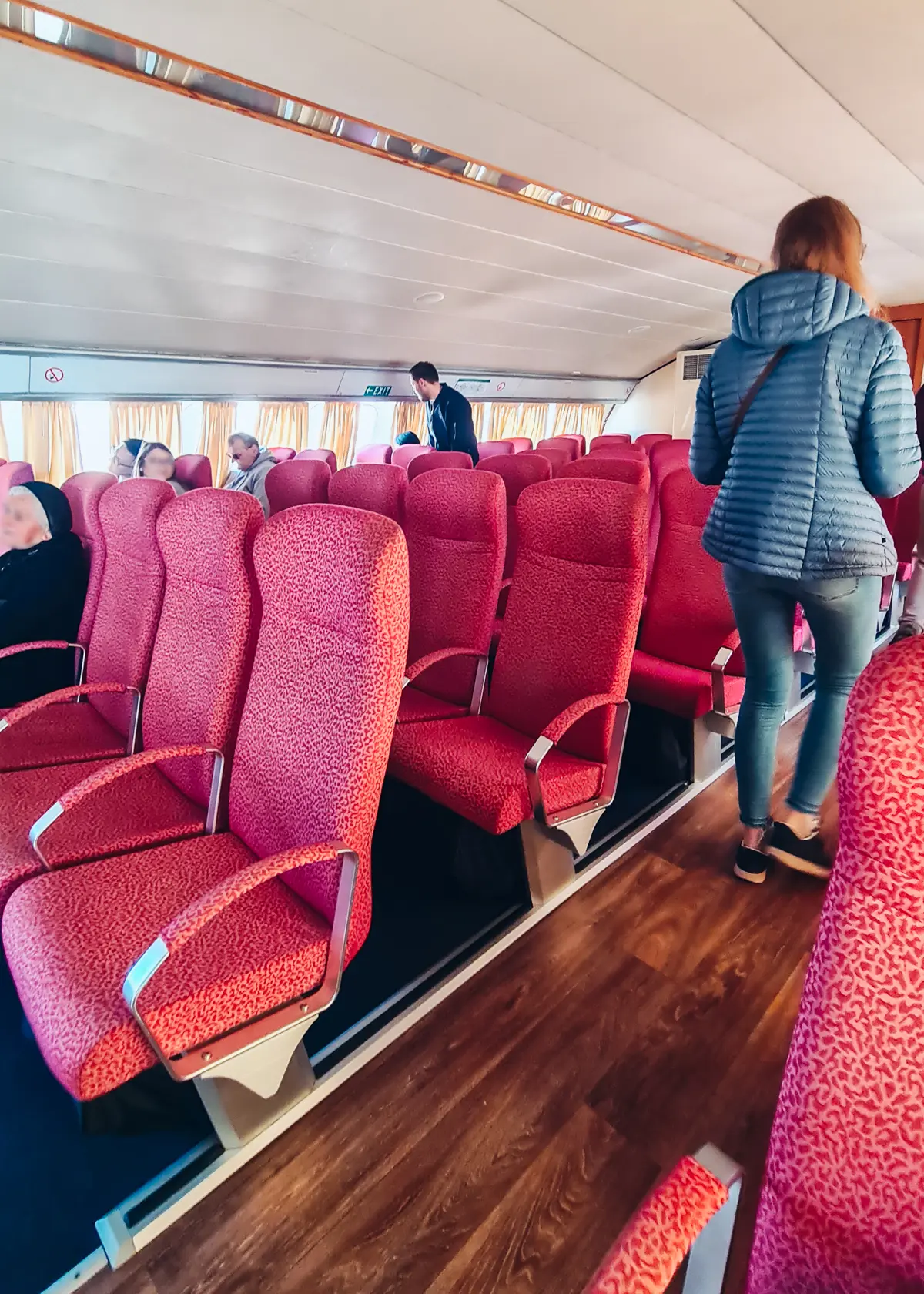 Woman wearing blue down jacket and jeans, walking between two rows of red seats on the Saranda to Corfu Ferry.