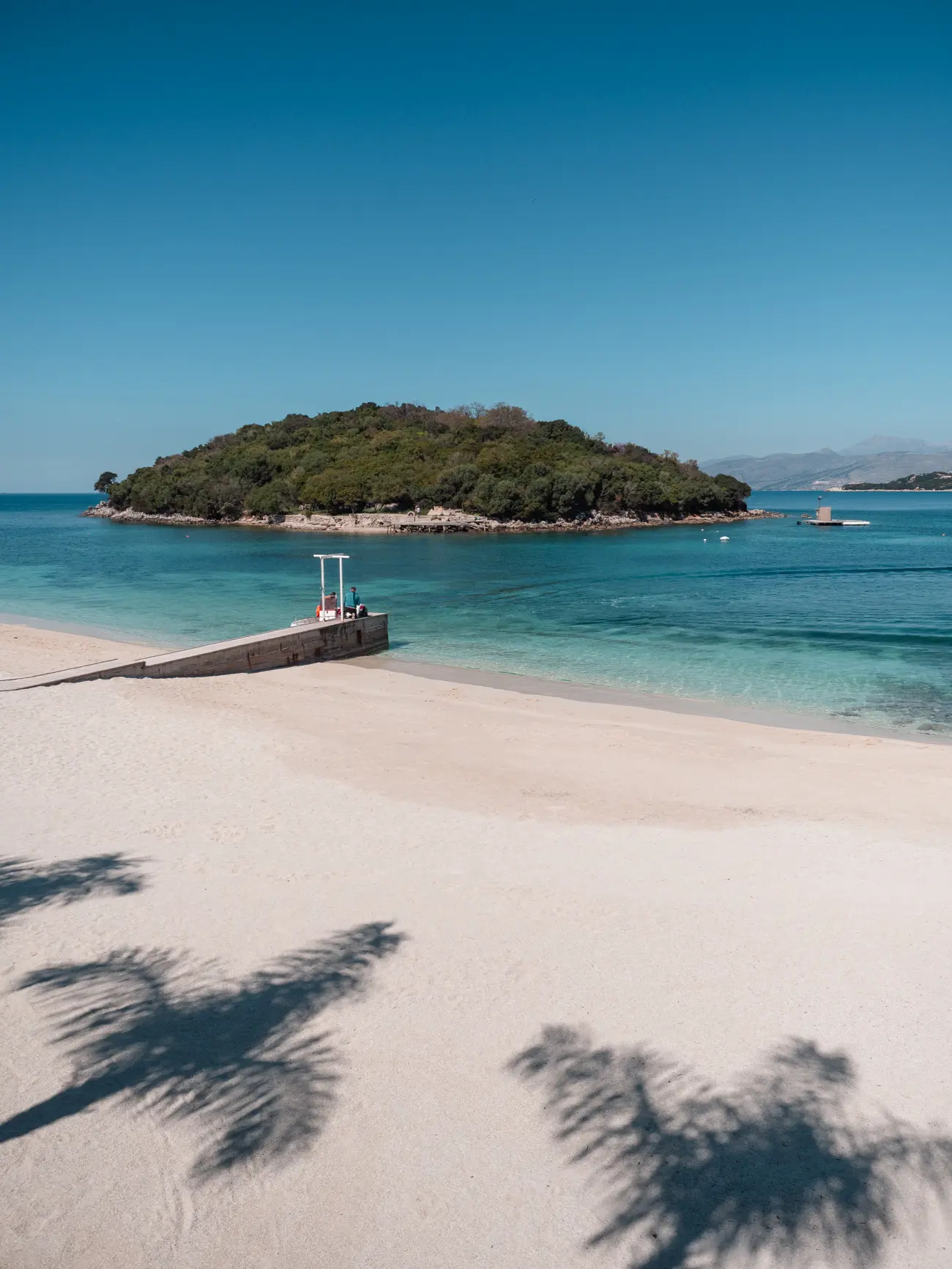 View of a narrow pier on the empty white-sand Ksamil Beach with an island in the background and two palm tress shadows on the sand.