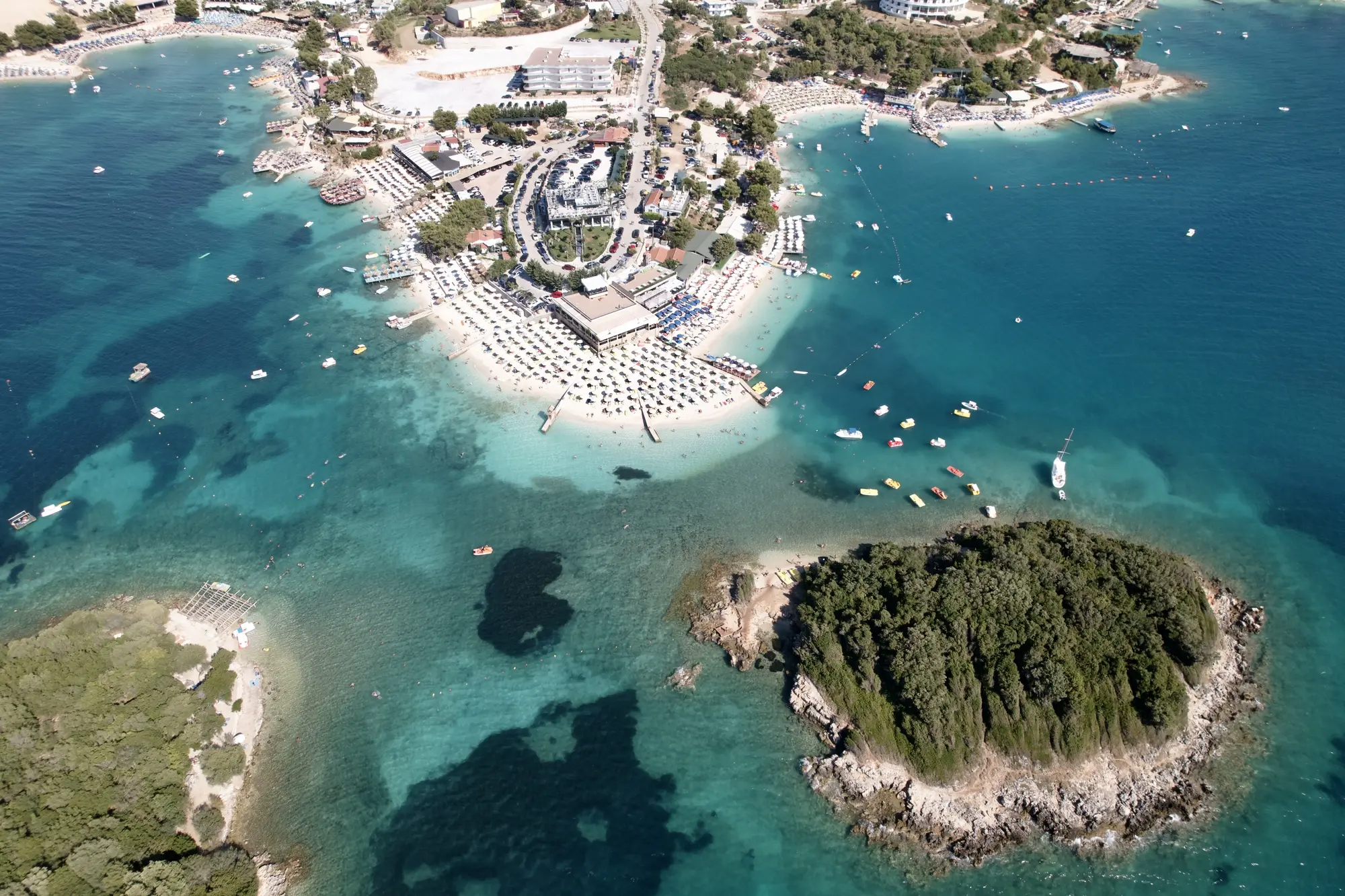 Aerial view of of the white-sand Ksamil Beach covered in sunbeds with smaller coves on both sides and two green islets in the turquoise ocean. A complete guide to Ksamil.