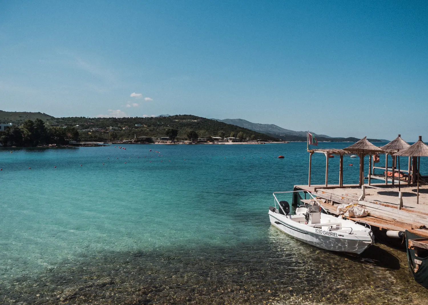 Small white boat docked next to a wooden deck with parasols above the turquoise ocean with a green island in the background in Ksamil.
