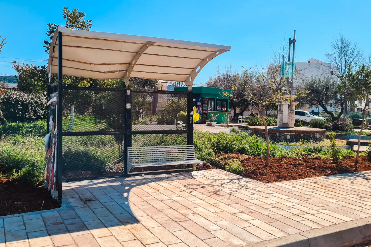 Black metal bus stop with glass sides and white roof, in front of a green garden on a sunny day in Ksamil Albania.