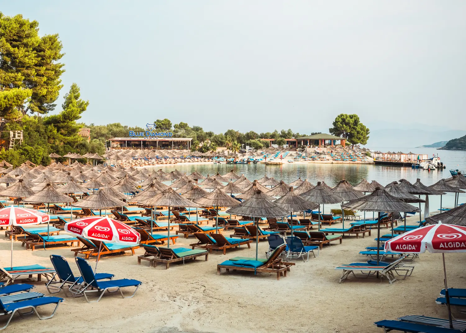 View of the light sand Lori Beach covered in blue sunbeds with straw umbrellas, with another beach club called Blue Diamond in the background, early in the morning in Ksamil.