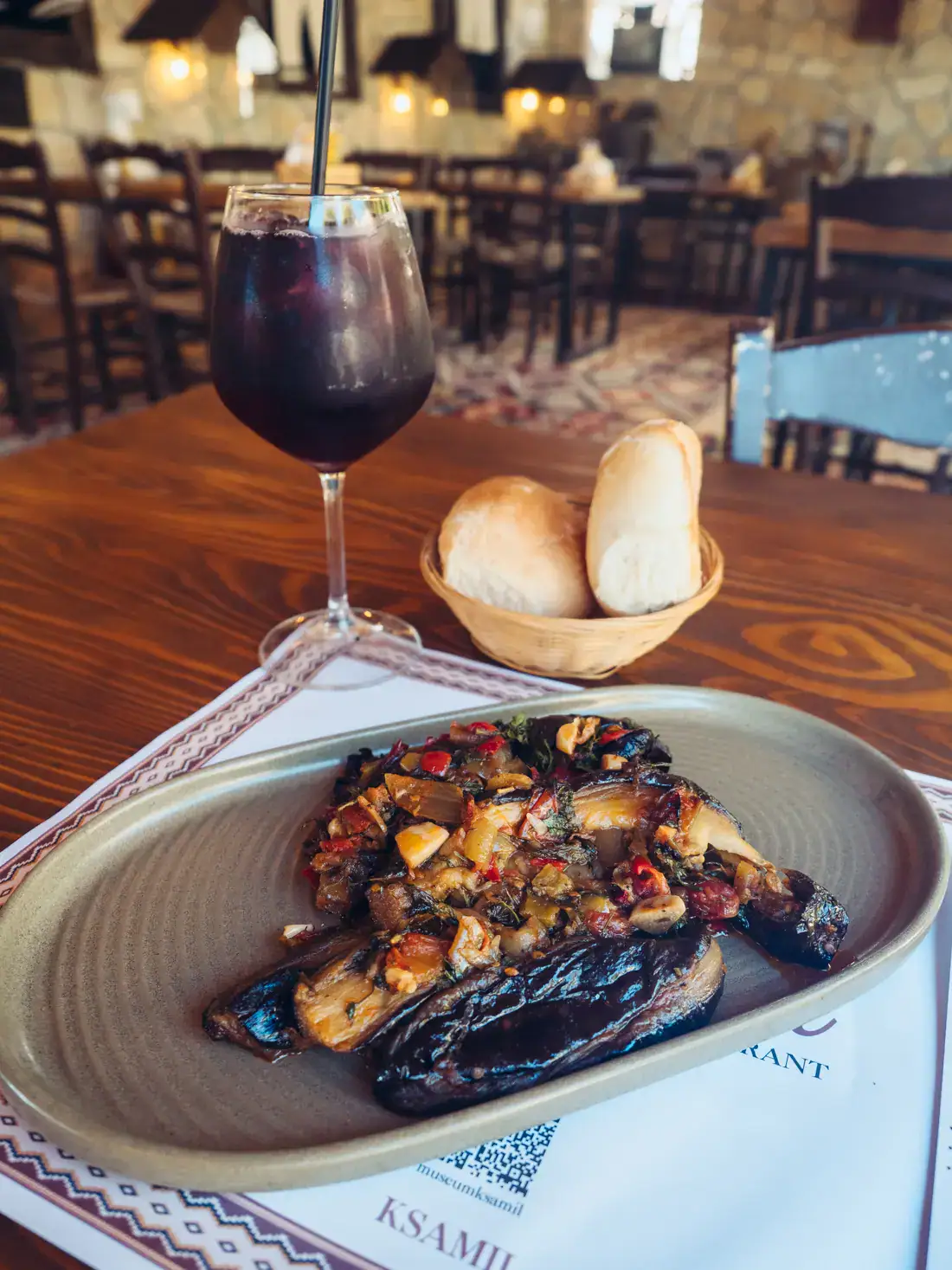 Rectangle grey plate with a baked eggplant, bread basket int he background and a large glass of delicious blueberry iced tea at Museum Restaurant, one of the best in Ksamil.