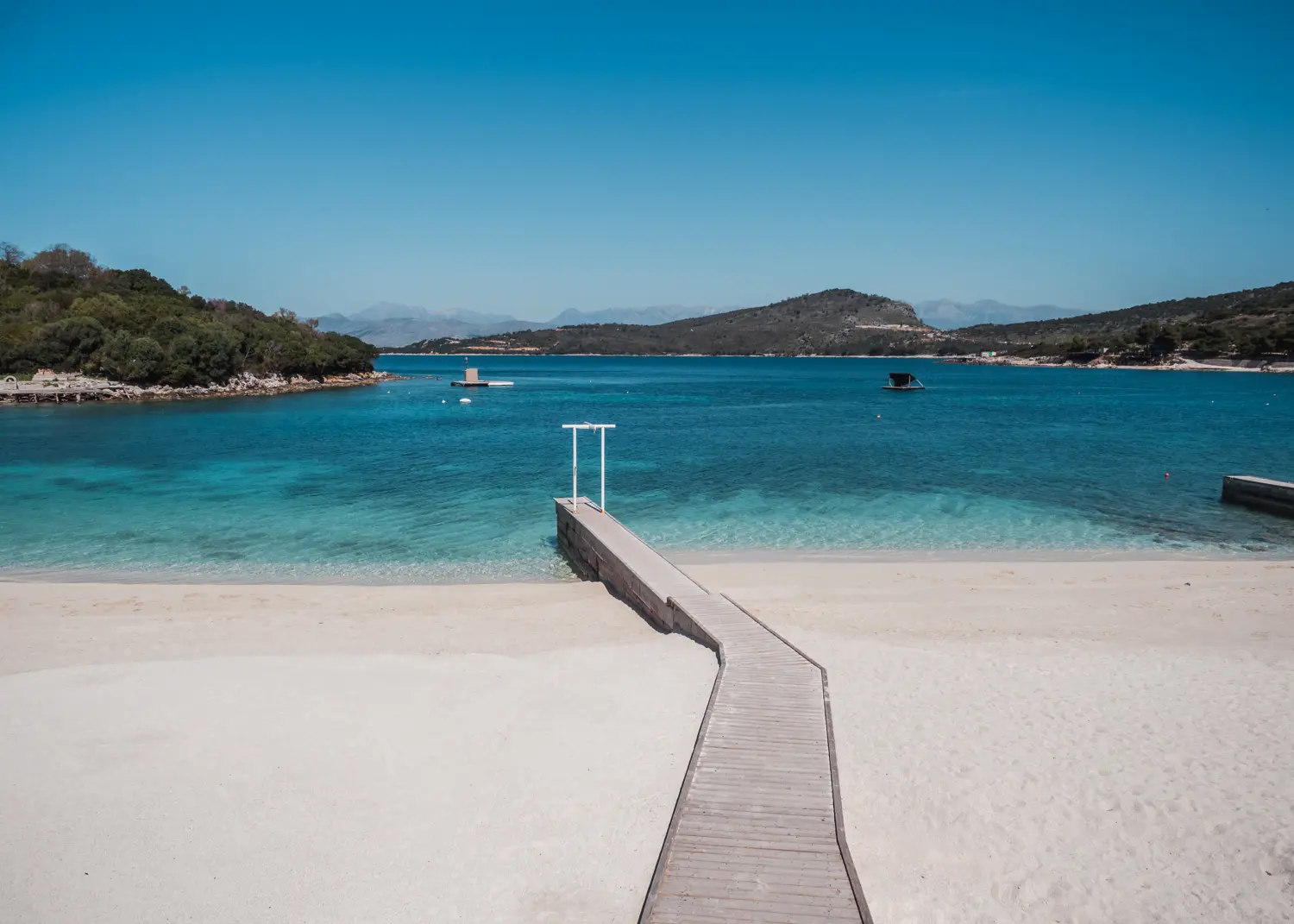 Narrow wooden pier with white sand on both sides, going in to the blue water with islands covered in greenery in the background on a beautiful sunny day in Ksamil, Albania guide.