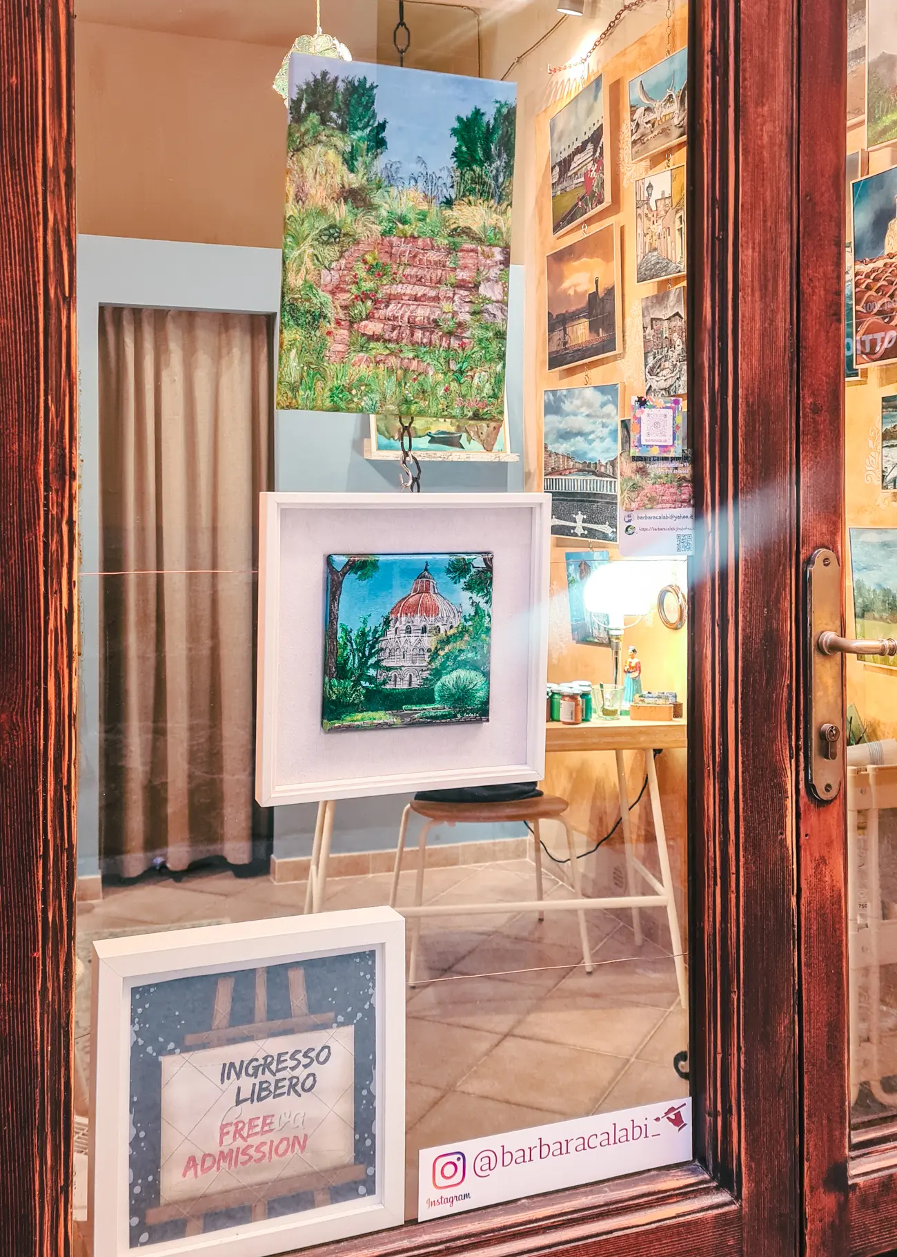 View of the window of a small gallery with two paintings of Pisa hanging and a large yellow wall with paintings inside in the background, seen during one day in Pisa.