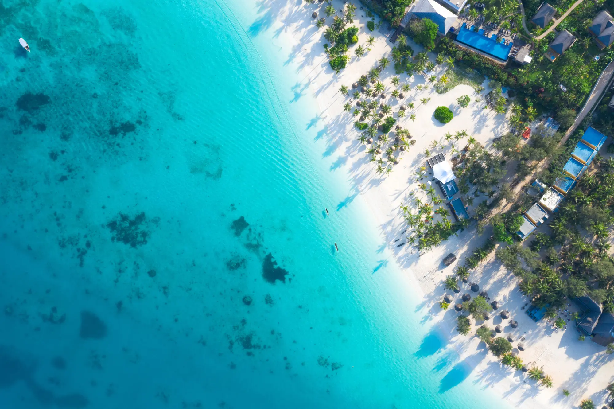 Aerial view of a bright white sand beach, clear turquoise ocean and a resort area with pools and green trees, how to book a Tanzania Zanzibar safari.