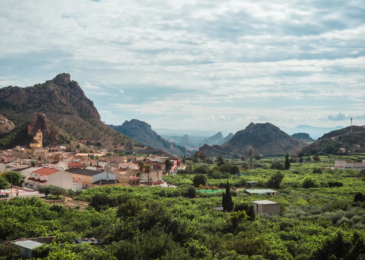 View of the small village of Ricote in Ricote Valley surrounded by lush fields and forest with mountains in the background on a cloudy day.