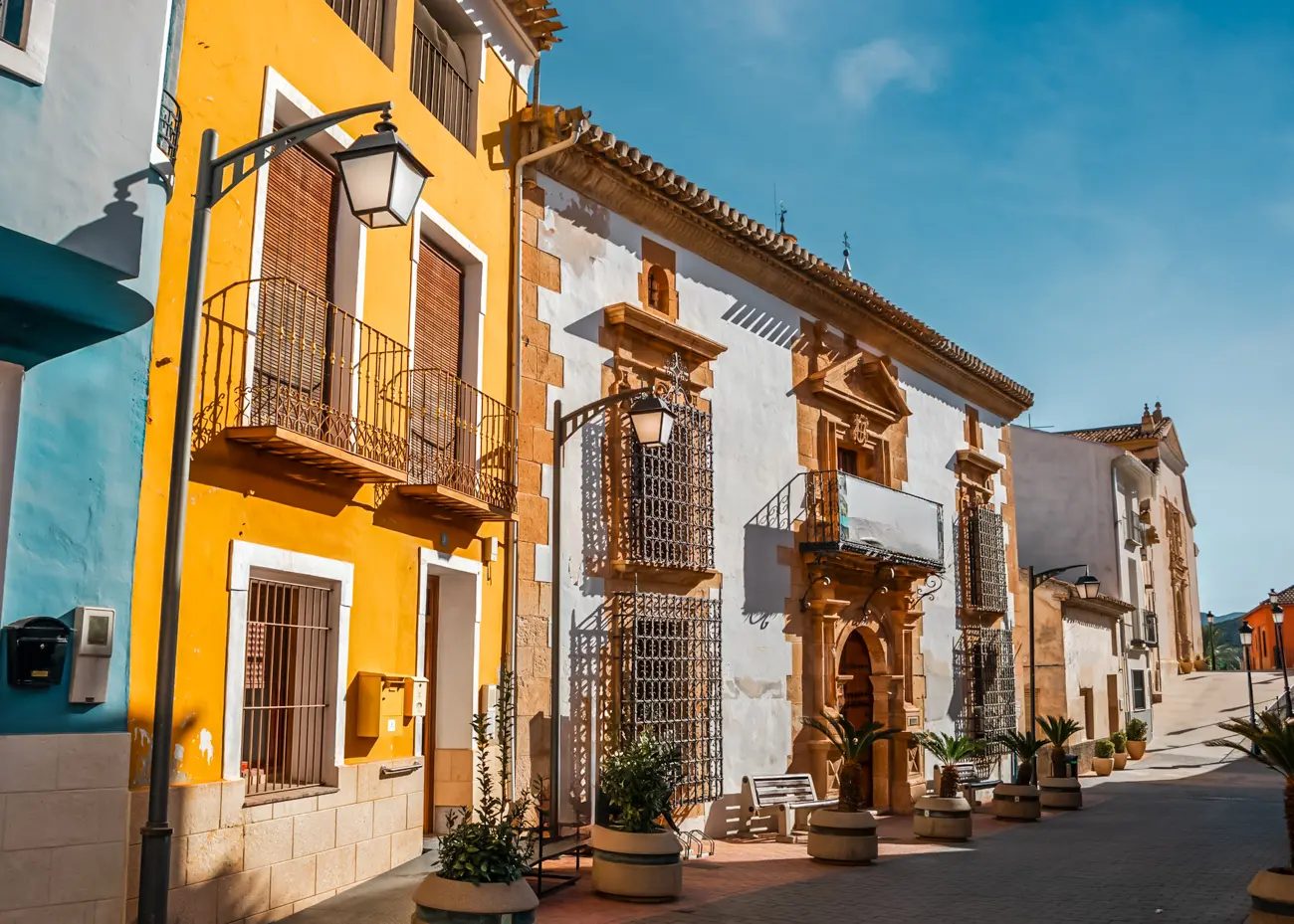 A street with a light blue, a yellow and a white house with brown details in Ricote, Ricote Valley.