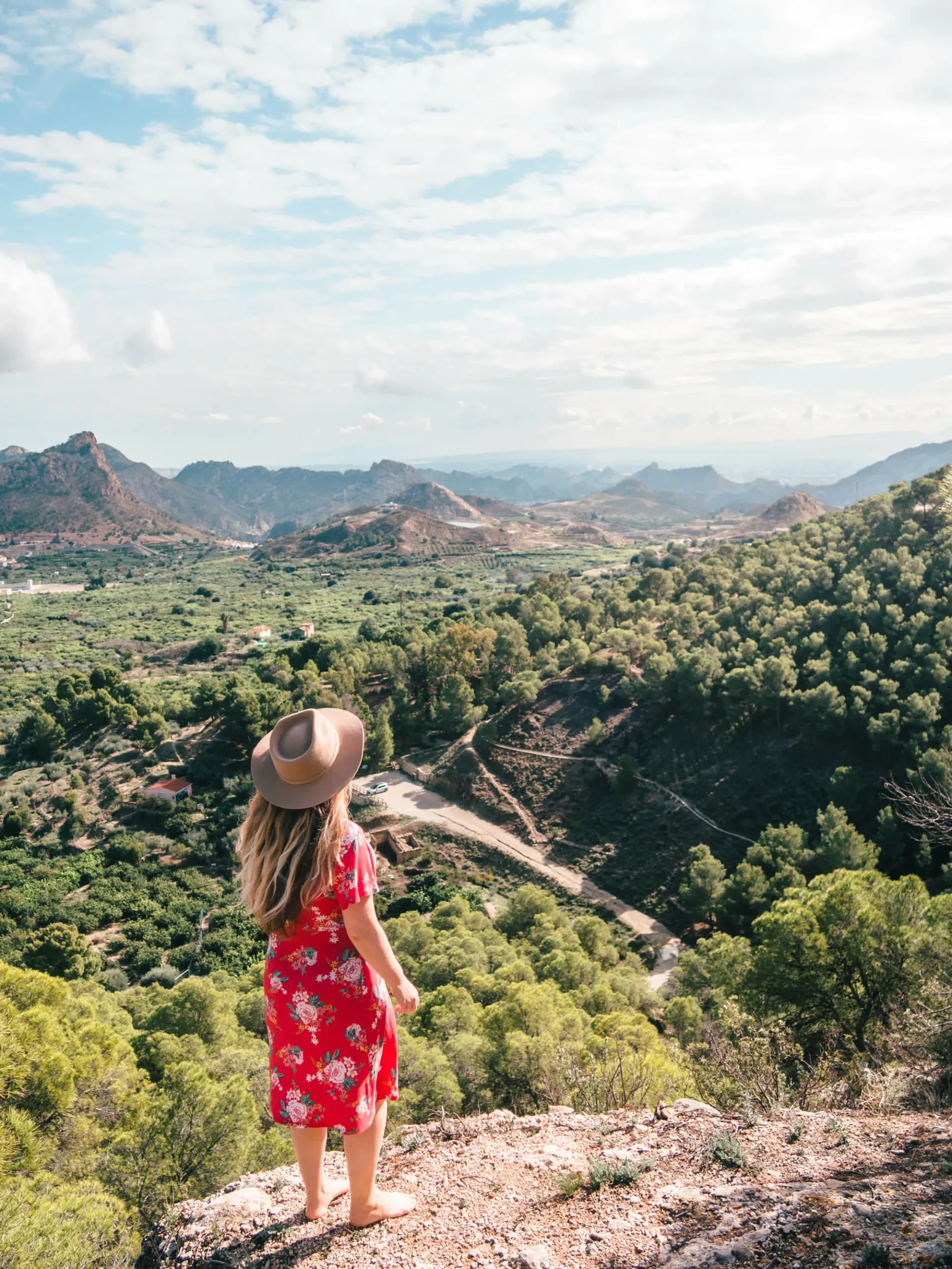 Woman with long hair, wearing a red dress and beige hat, standing on a cliff edge looking out over the lush peaks of Ricote Valley from Mirador del Cabecico de la Doctrina viewpoint.