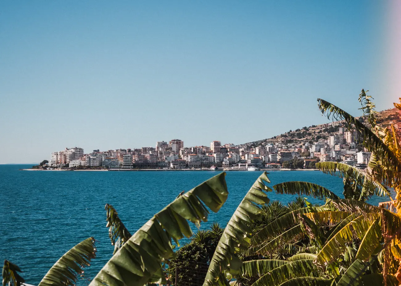 View over palm leaves of the town of Saranda with many high-rise buildings along the blue ocean on a sunny day.