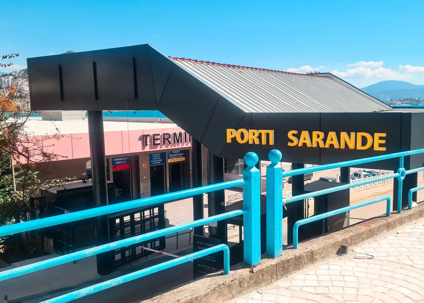 Light blue metal fence in front of a black roof with yellow text saying Port Sarande, in front of the terminal building, taking the Saranda to Corfu Ferry.
