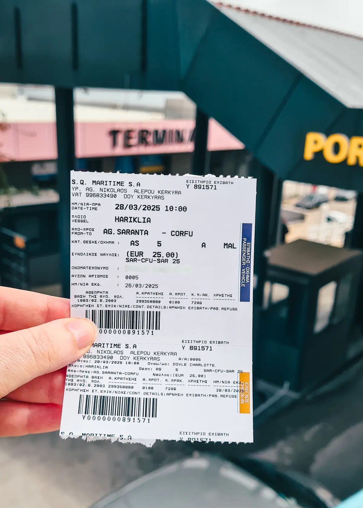 Close up of hand holding a white ticket with small black text in font of the black and salmon colored ferry port in Sarande, before taking the ferry to Corfu.