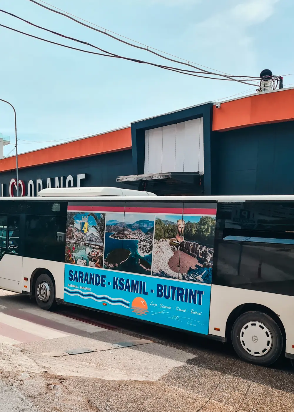 White bus with blue "Saranda-Ksamil-Butrint" sign on the side, driving past a black and orange building in Saranda on its way to Ksamil, Albania.