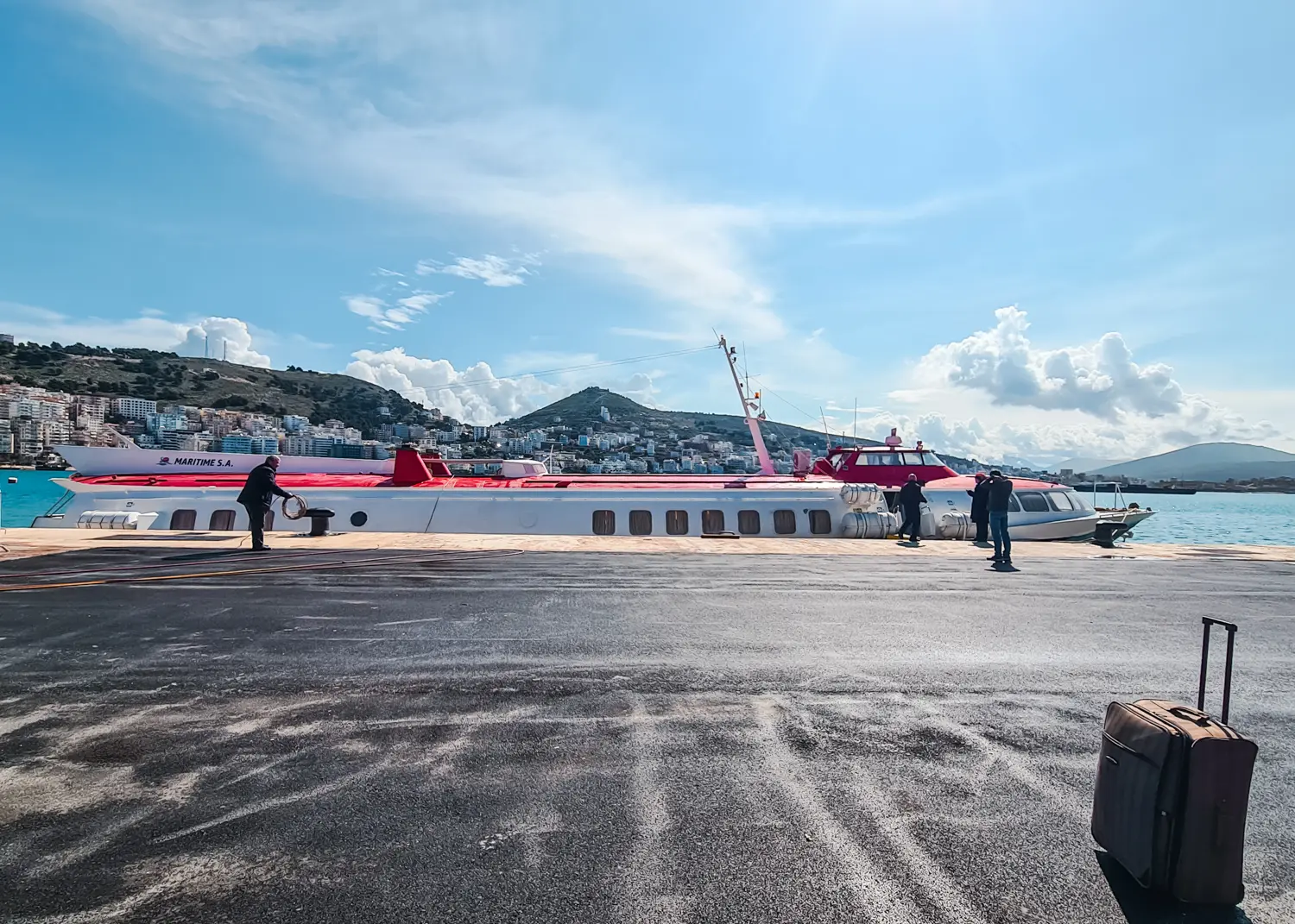 Large asphalt waiting area at Sarande ferry port with one black suitcase in the right corner, looking at the docked red and white Finikas Lines ferry to Corfu, on a sunny day.