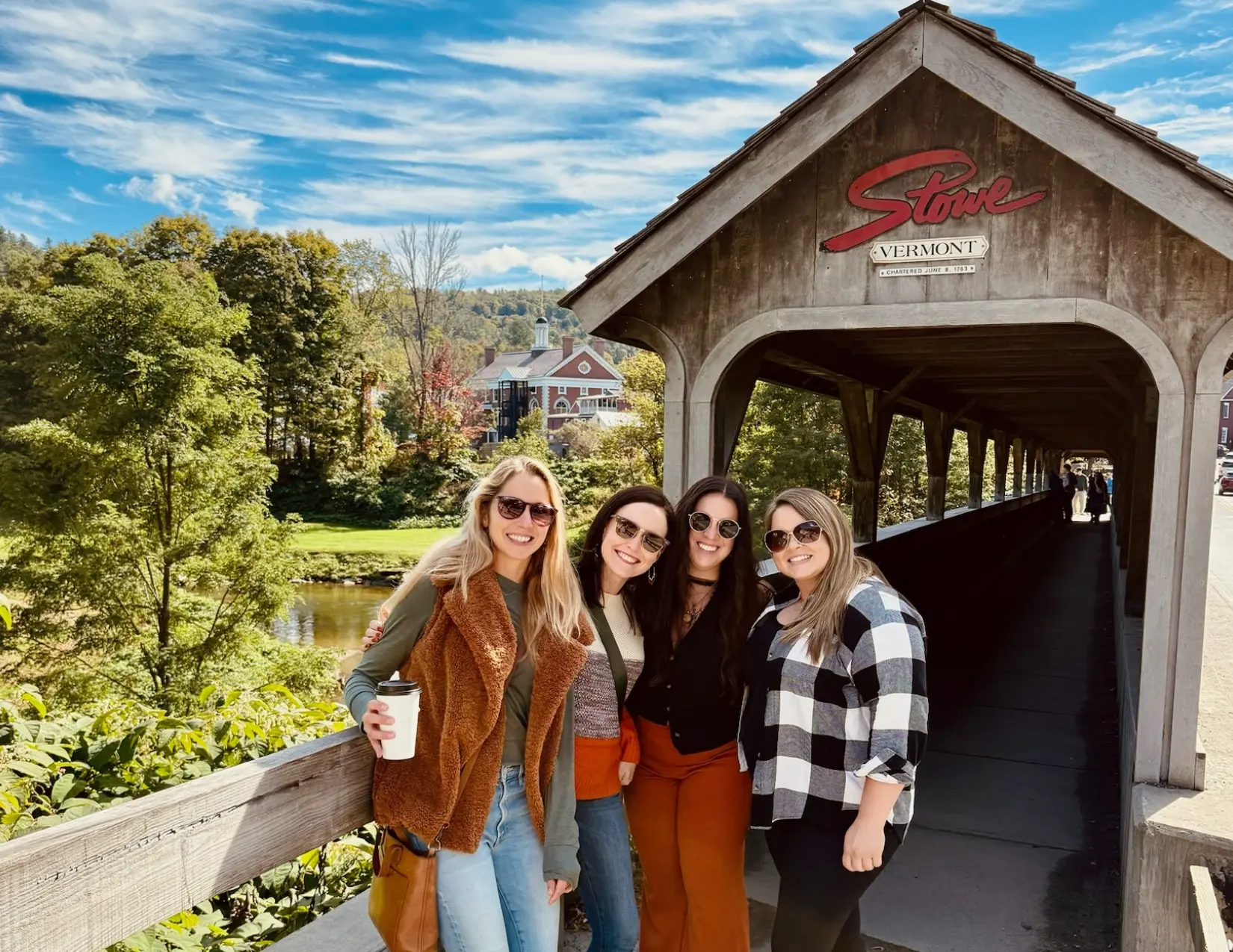 Four girls in fall outfits and sunglasses standing in front of a covered part of the Stowe Recreation Path in Vermont.