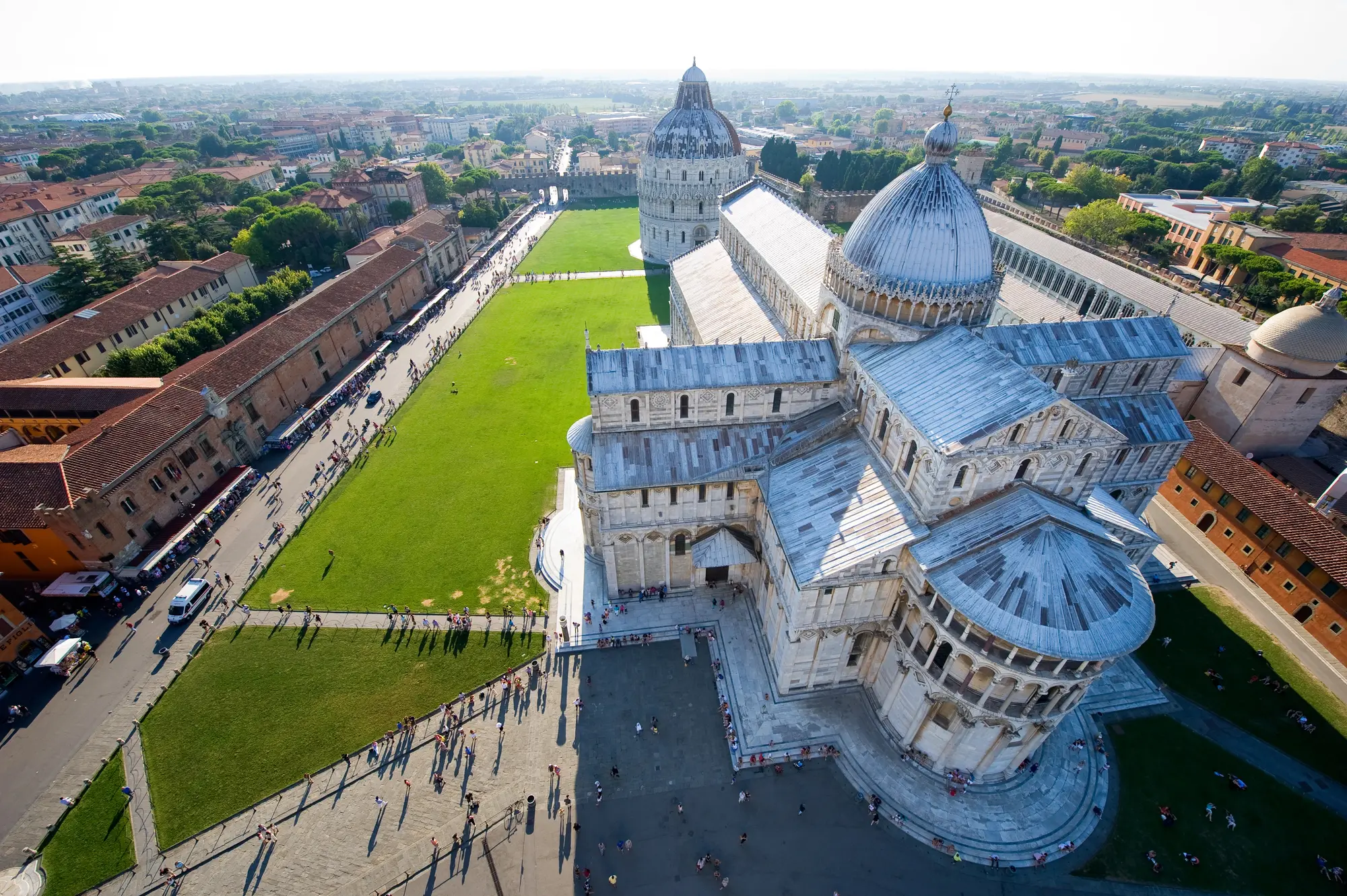 View from the Leaning Tower of Pisa of Piazza dei Miracoli with the light grey/beige Pisa Cathedral and Baptistery with a long green lawn in the right.