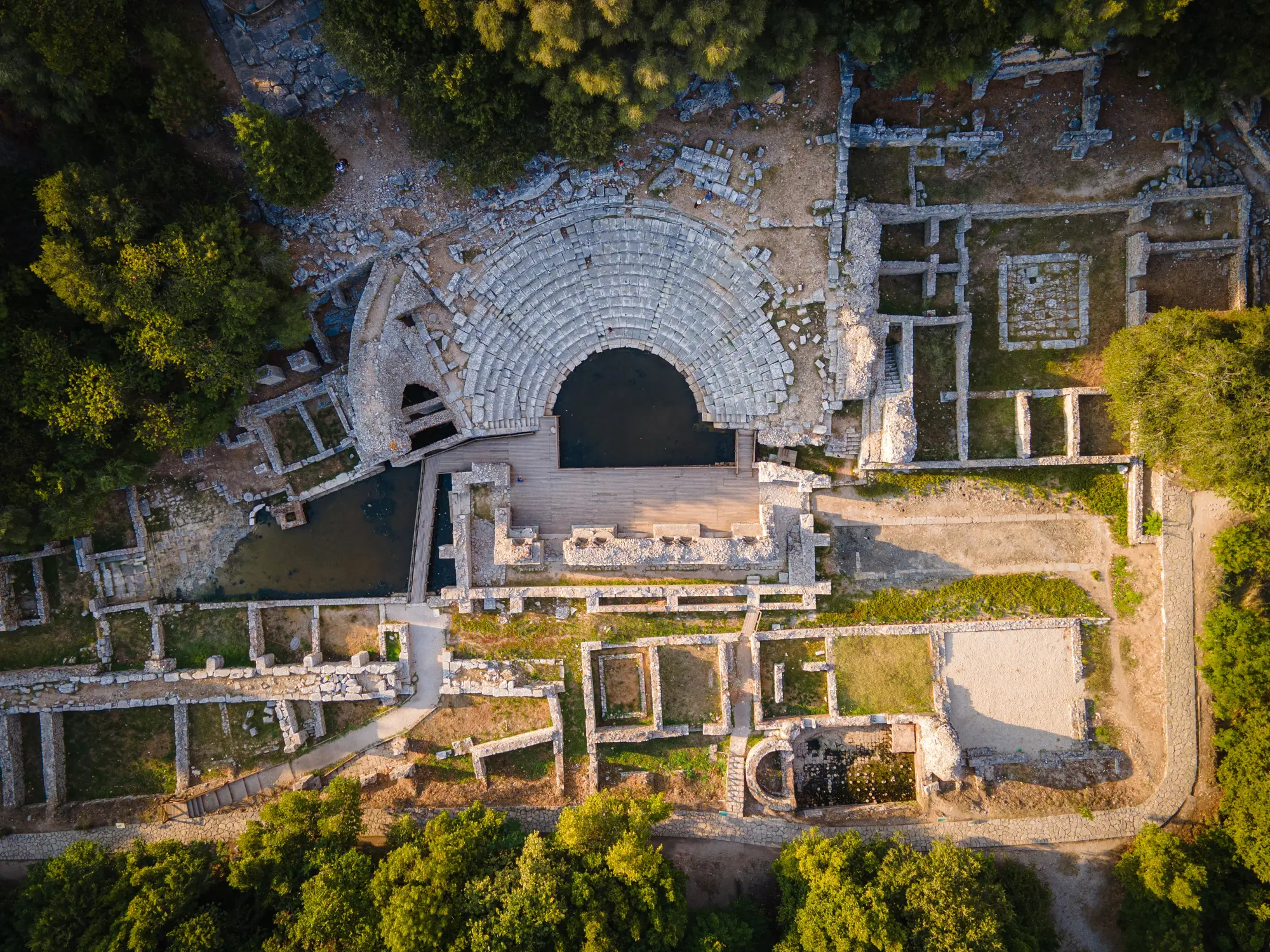 Aerial view of the old grey stone amphitheatre in Butrint National Park surrounded by other ruin buildings and lush greenery in the morning light, one of the top things to do while in Ksamil, Albania.
