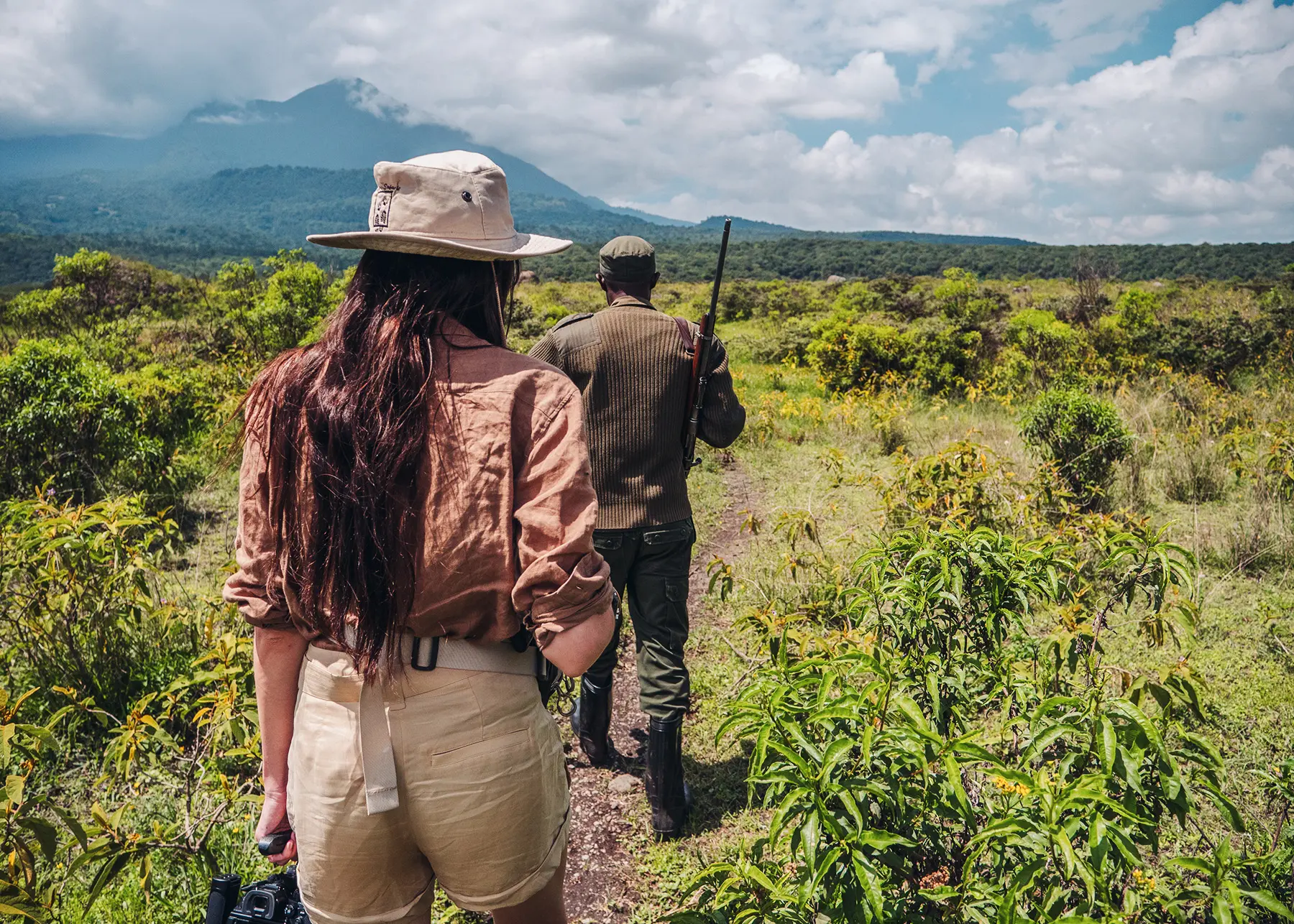 Woman with long brown hair, wearing a light brown shirt, beige hat and beige shorts, walking after a safari guide in a lush green landscape during a Tanzania Zanzibar safari.