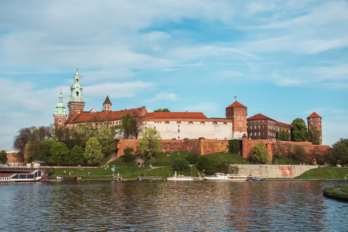 View of the majestic Wawel Castle, made from red brick with three spires on the left and two towers on the right, seen from the river, surrounded by green grass and trees. Where to stay in Krakow.