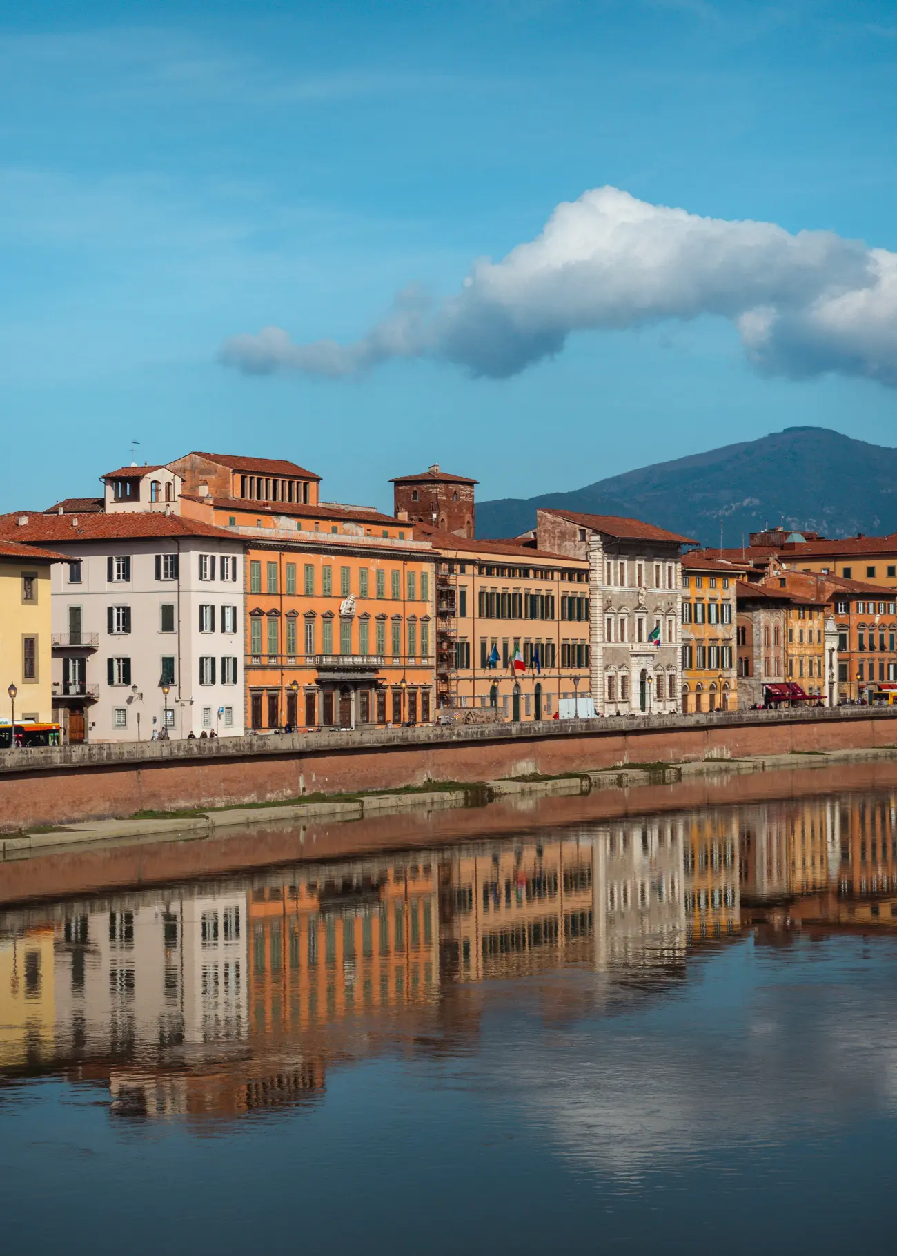 Light beige, orange, white and yellow houses reflecting in the calm Arno River on a sunny day with some clouds in Pisa.
