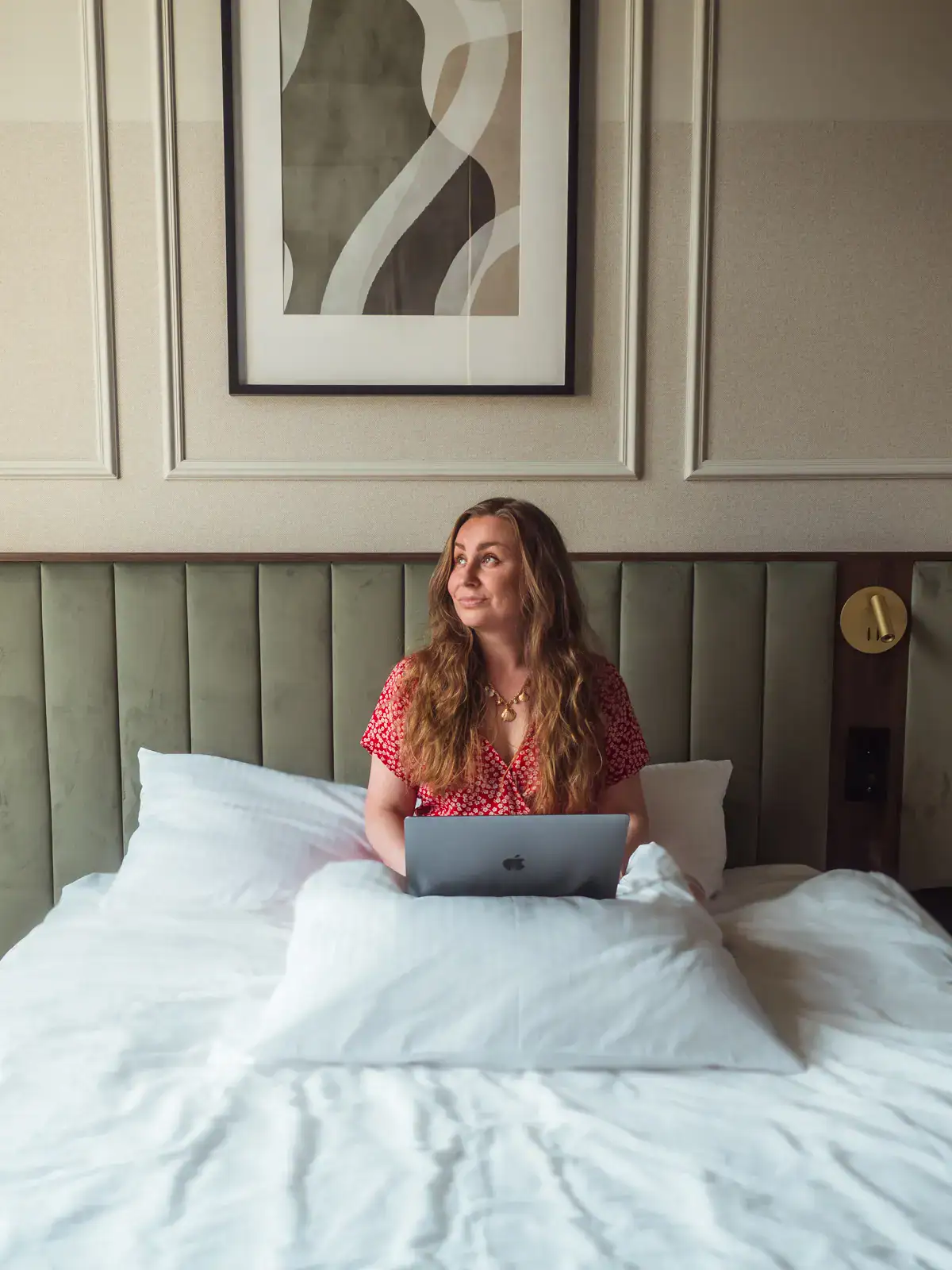 Woman in a red dress with long light brown hair, sitting in a bed working on her computer, green headboard, one of the best apartments to rent in Krakow.