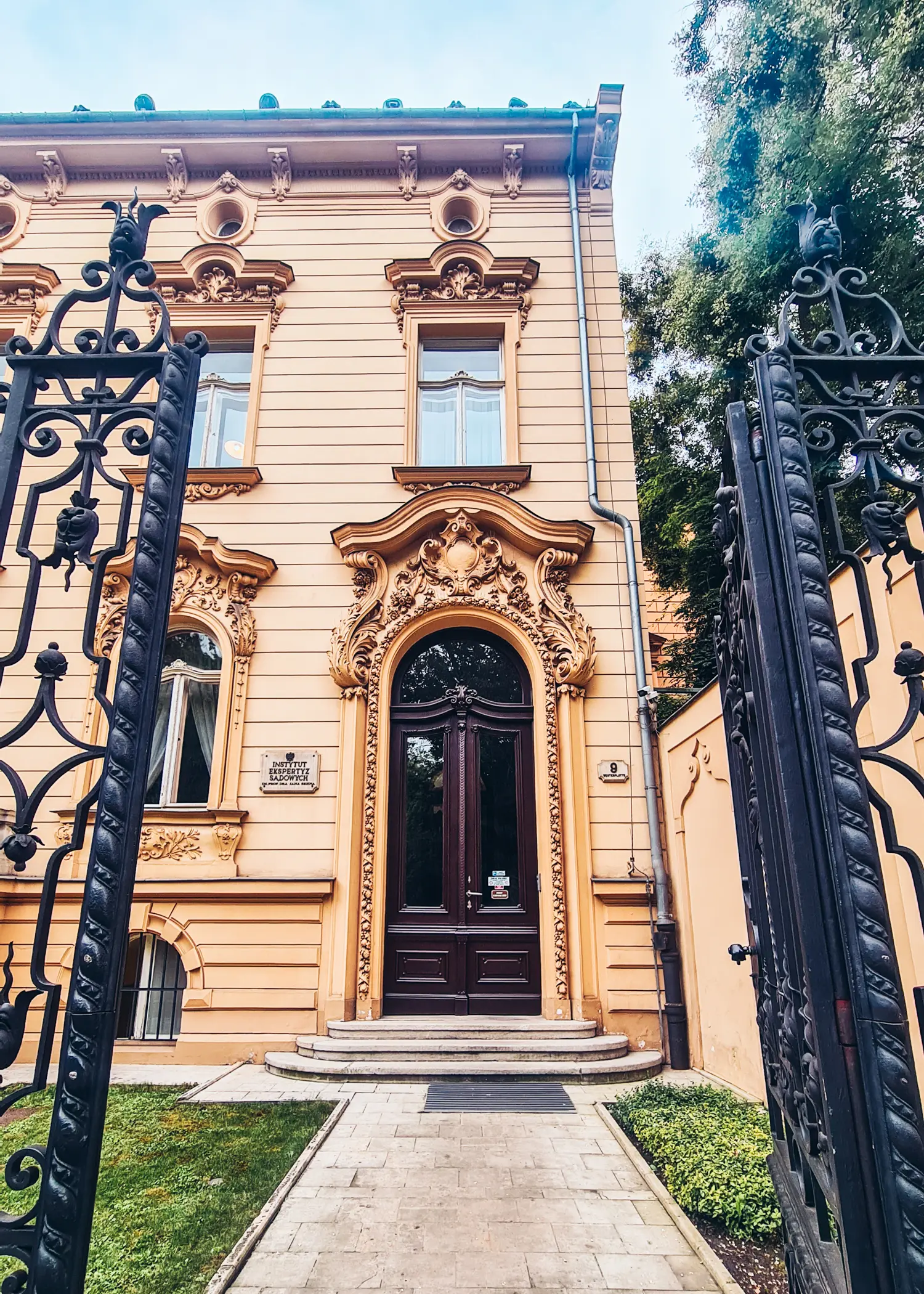 Looking through an ornate rod iron gate on a light yellow building with details around the brown, arched door and windows in the Wesola neighborhood, where to stay in Krakow.