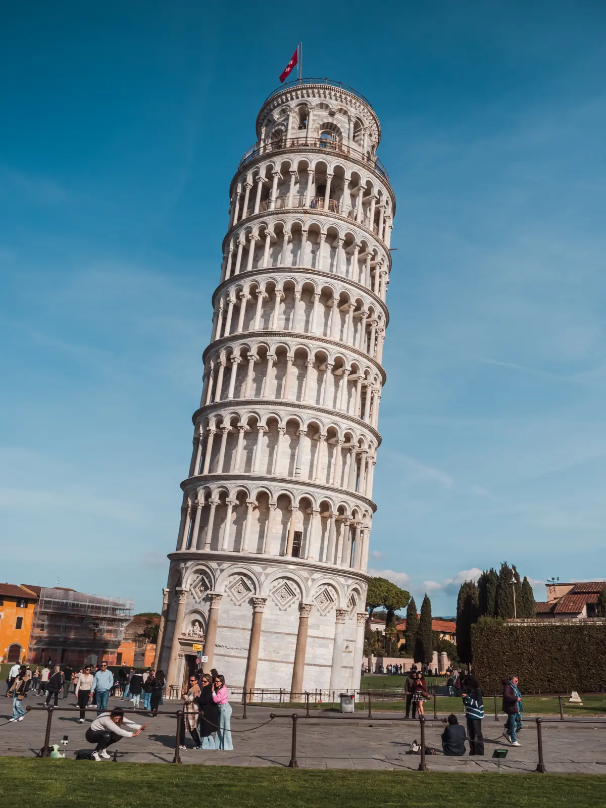 View of people taking the typical tourist photos in front of the Leaning Tower of Pisa with many arches made from white marble and limestone with a red flag on top, set against a deep blue sky.