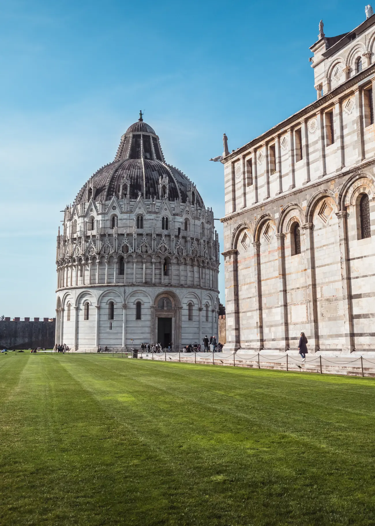 The round, ornate Baptistery on Piazza dei Miracoli, behind Pisa Cathedral with a bright green lawn in front on a sunny day.