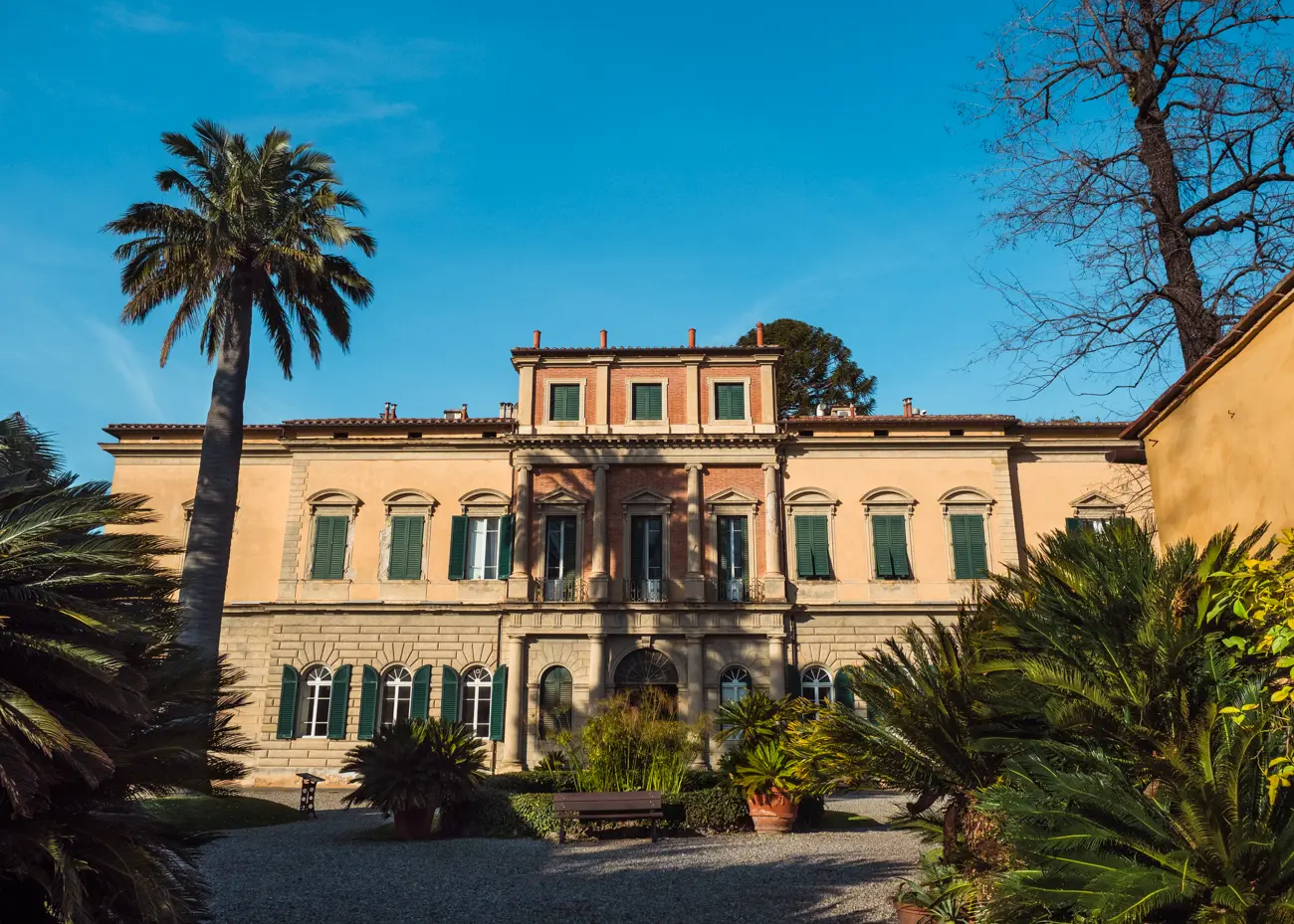 Large light beige/orange manor house with a tall palm tree on the left and green bushes on both sides in Pisa Botanical Garden, the highlight of my one day in Pisa.