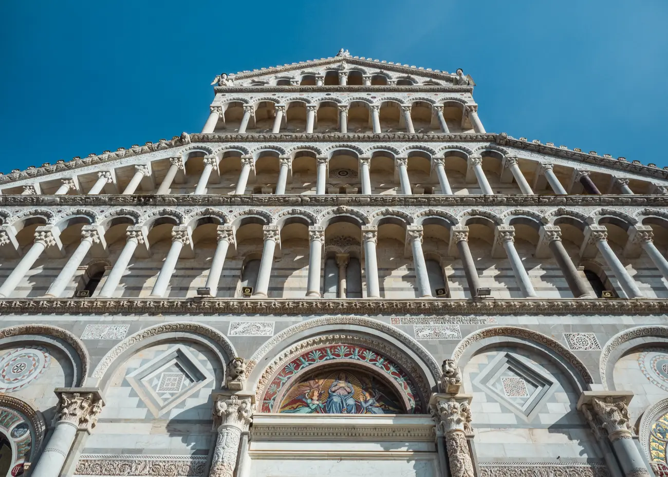 Looking up at the majestic Pisa Cathedral with arches going up the wall and a colorful fresco in the middle on a sunny day with blue skies.