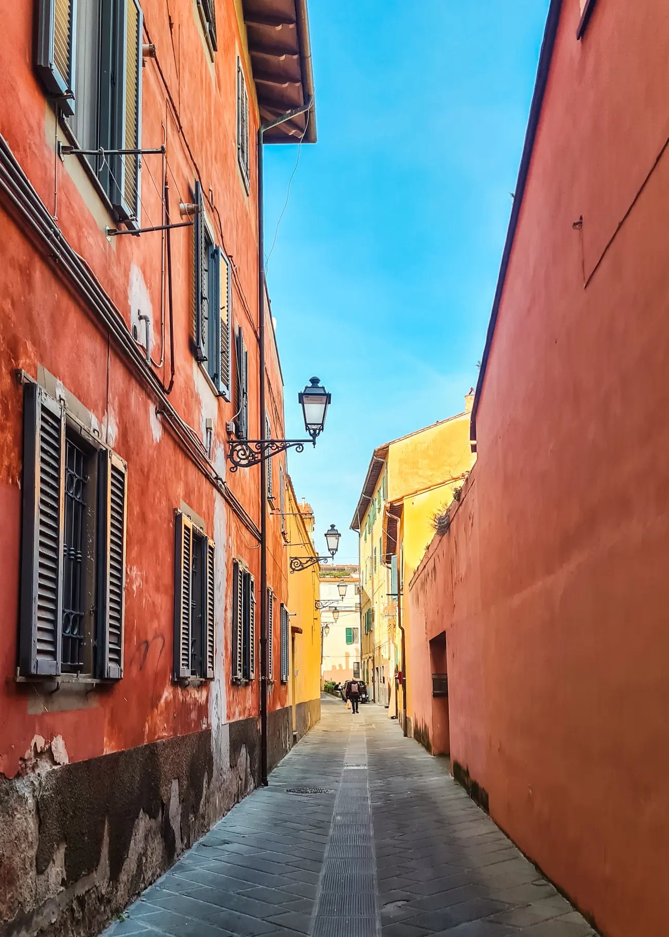 View of a narrow lane between two orange houses, the left with brown shutters and a light, yellow houses further down, on a sunny day in Pisa, Italy.