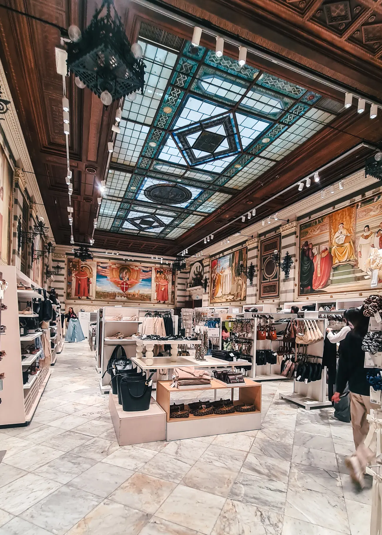 View of the accessories section in an H&M store in a grand old building with glass roof lined by dark wood in Pisa.