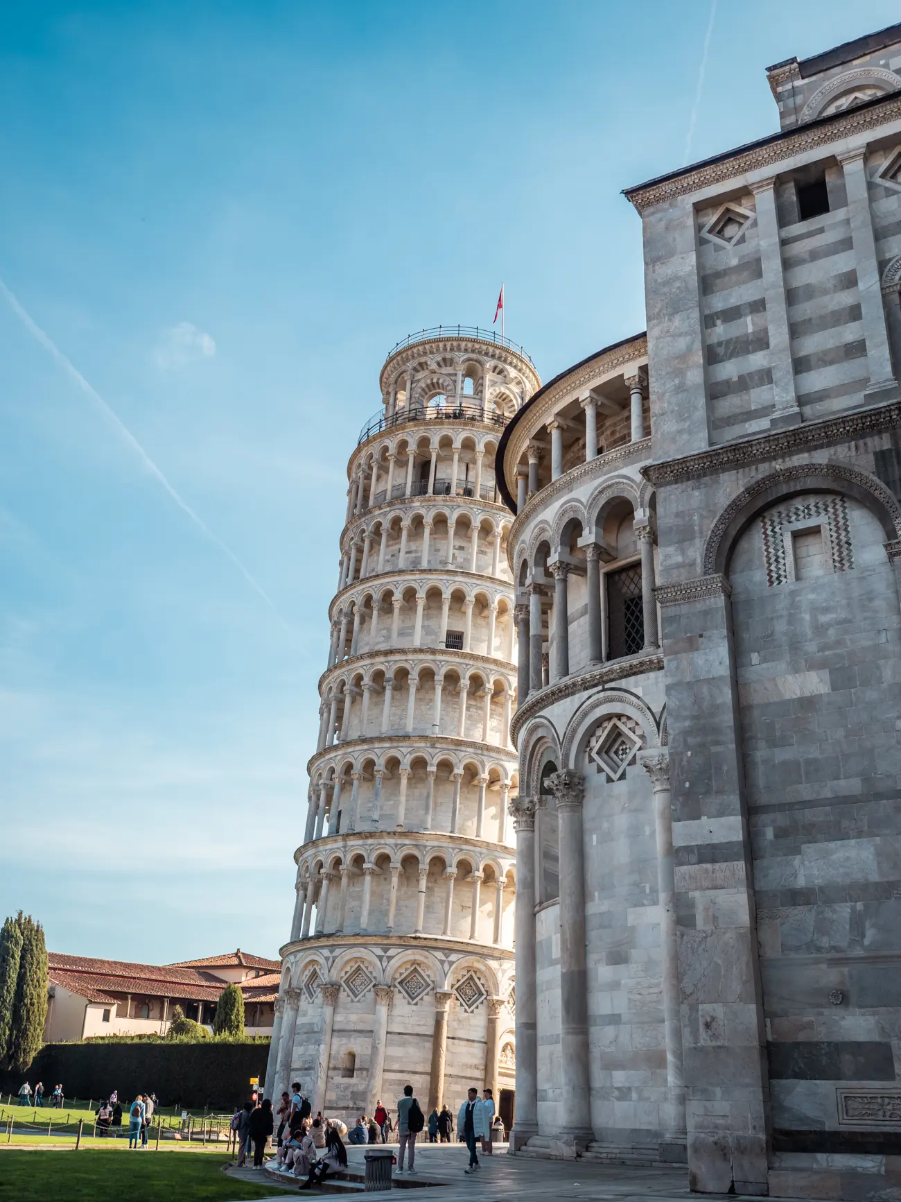 The Leaning Tower of Pisa made from white marble and sandstone seen from behind the majestic Pisa Cathedral made from grey stone, with people looking up at it on a sunny day in Pisa.