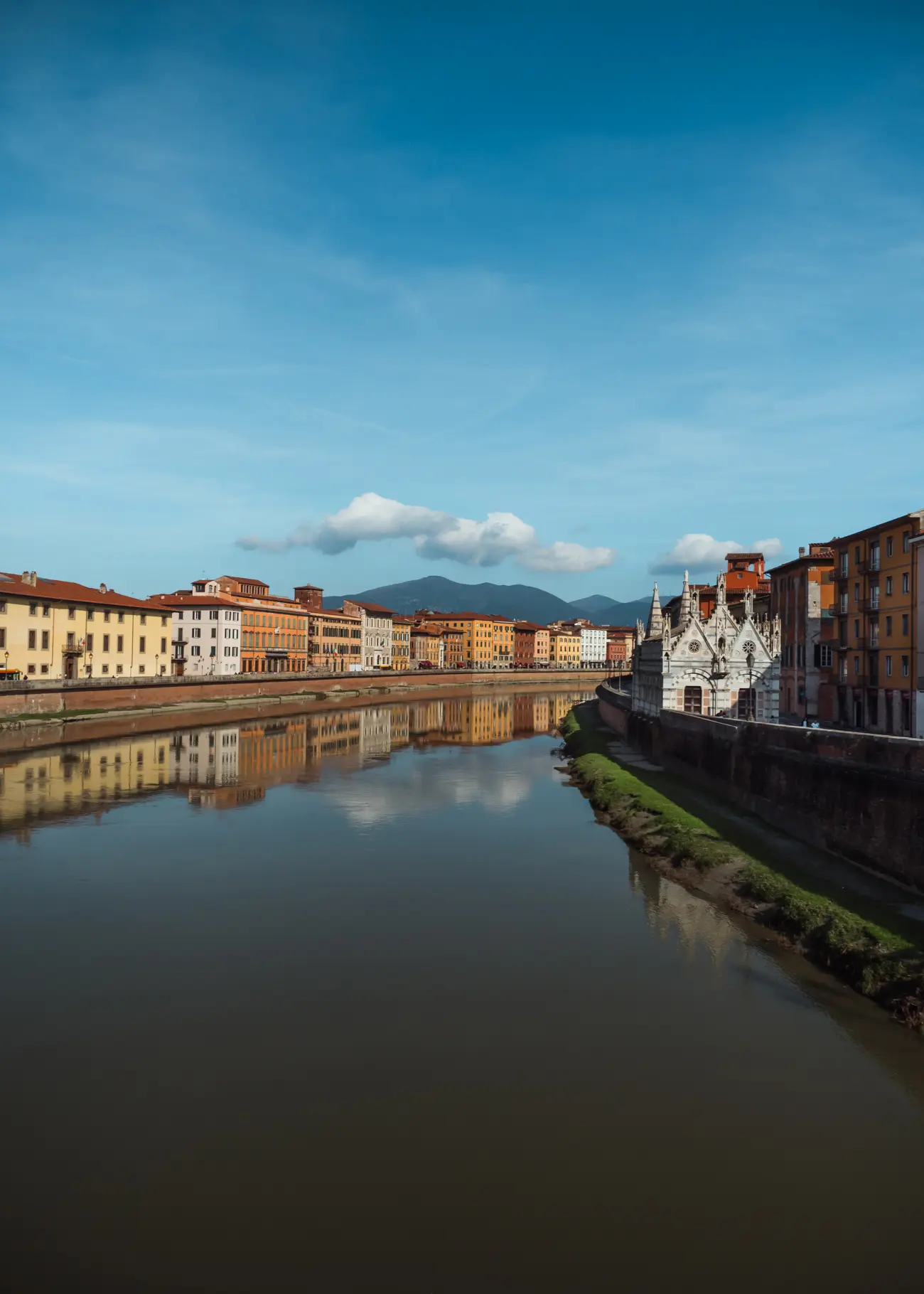 Light beige, yellow and orange houses reflecting in the calm Arno River on a sunny day in Pisa. Santa Maria della Spina, a tiny but gorgeous Gothic church stands on the right riverbank.