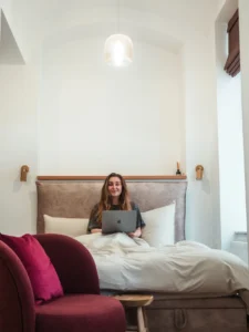 Woman with long dark blonde hair sitting in a bed with beige bedding with a laptop on her lap under a white arched roof in an apartment in Kazimierz, Krakow.