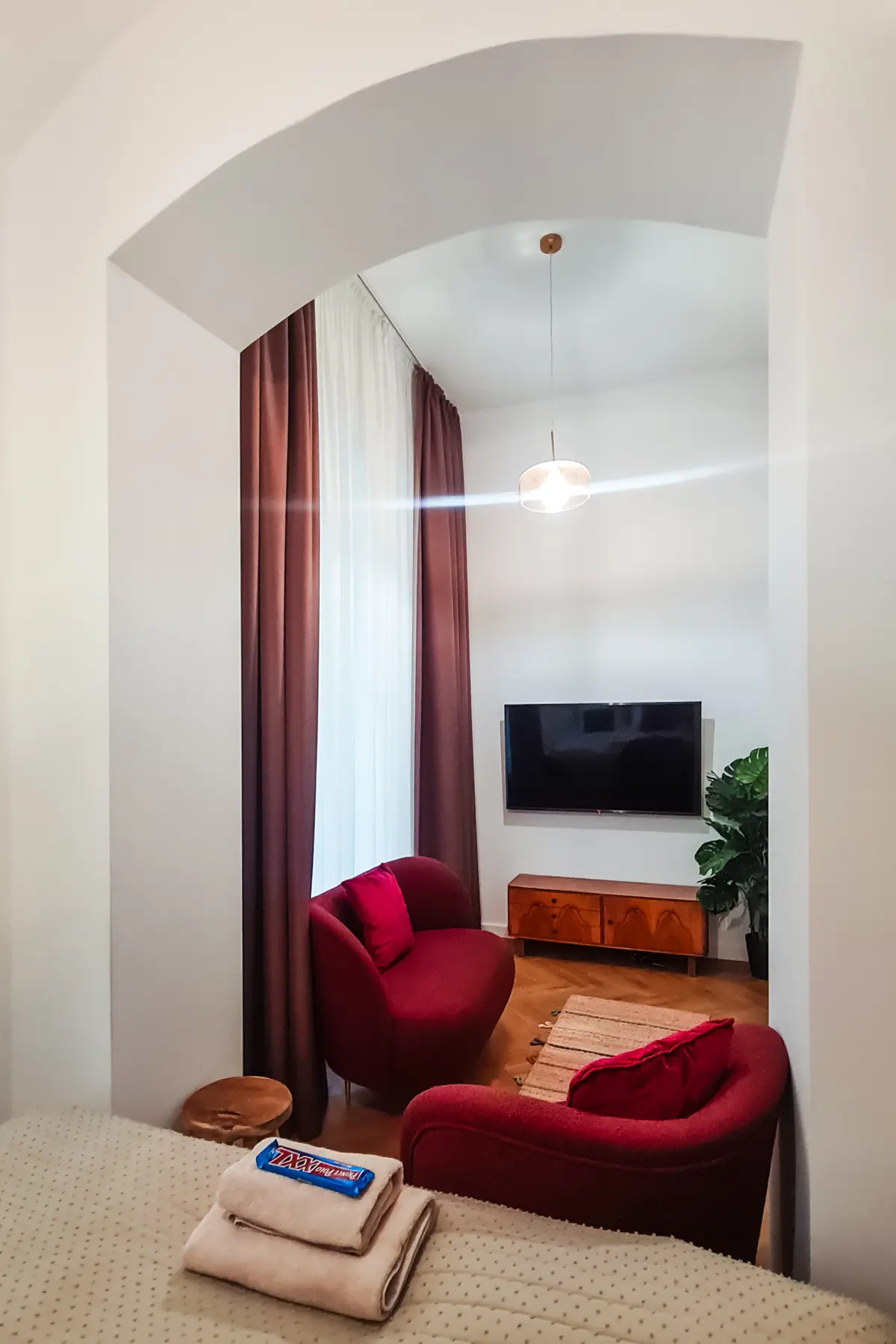 Looking through a white arch into a small living room with tall ceiling, red couch and chair and burgundy curtains in a beautiful apartment in Kazimierz, one of the best places to stay in Krakow.