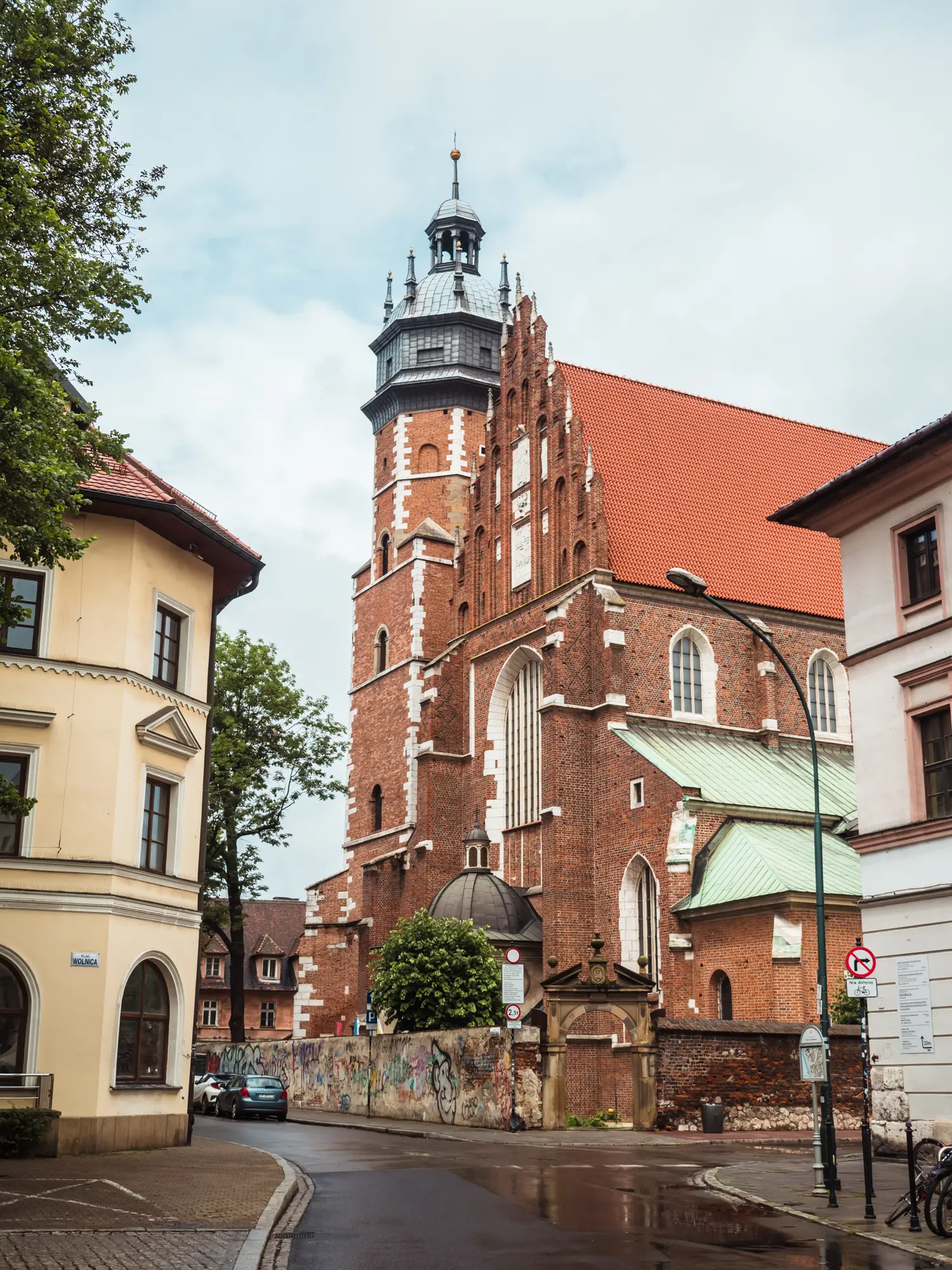 Tall red brick church with a spire and orange roof seen between two older buildings on a cloudy day in Kazimierz, one of the best areas to stay in Krakow.