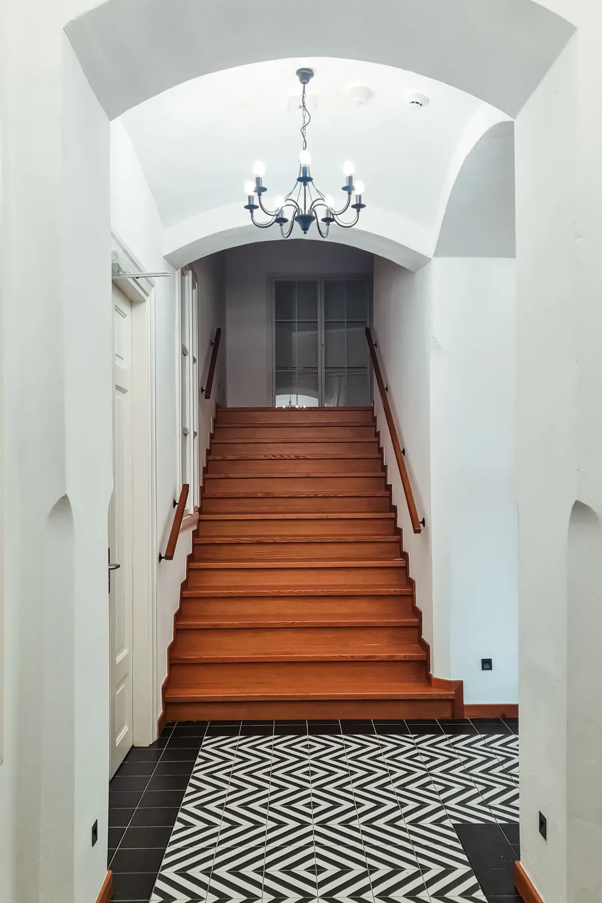 Black and white patterned floor and a wooden staircase going up under a white arch with a chandelier in an apartment building in Kazimierz, where to stay in Krakow.