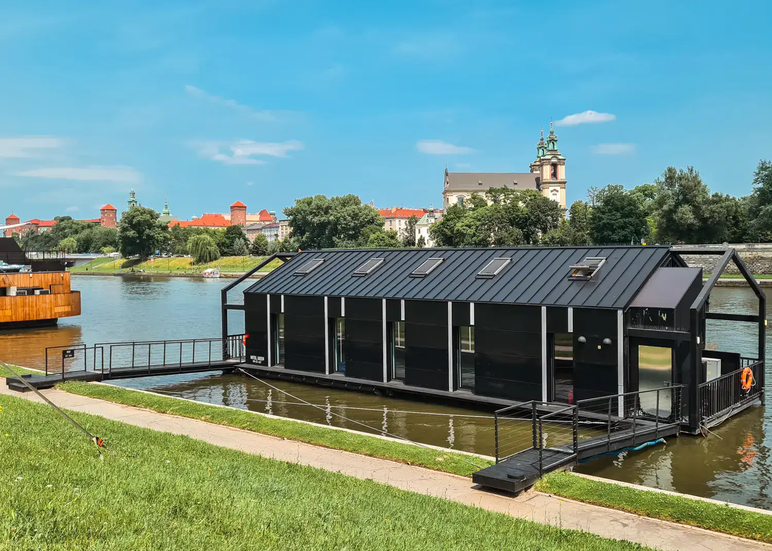 Black New Port Hotel building with 5 rooms on the river, two bridges connects it to the green, grassy river bank, with Wawel Castle and a church on a sunny day, where to stay in Krakow.