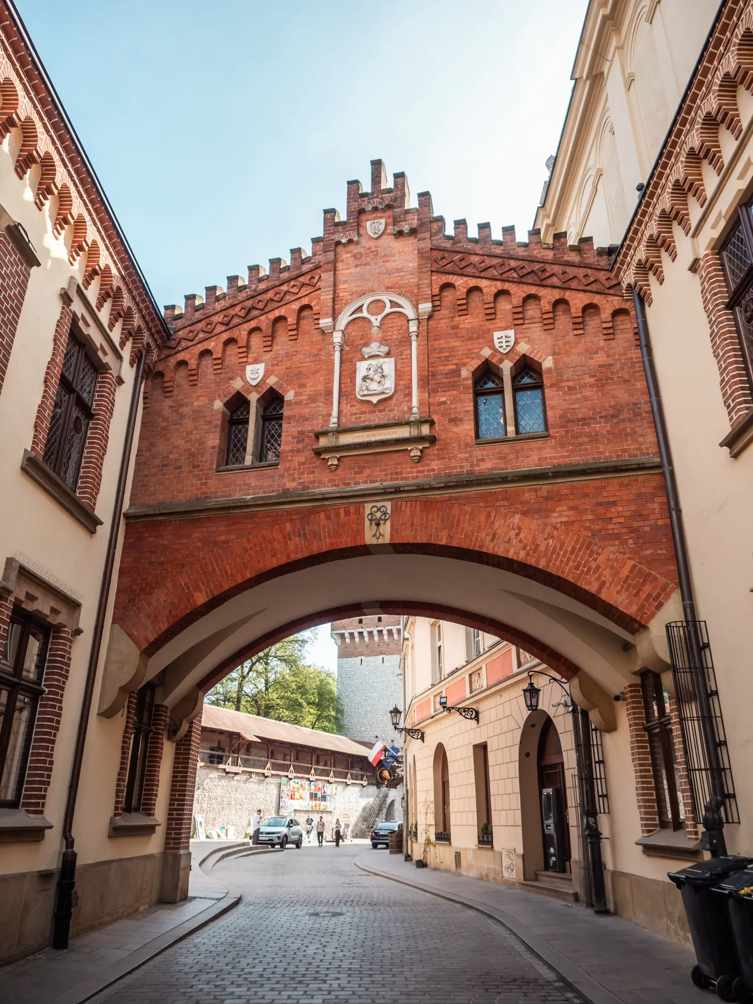 Red brick Medieval looking archway over a cobbled street between two buildings on a sunny day in Krakow Old Town, one of the best places to stay.