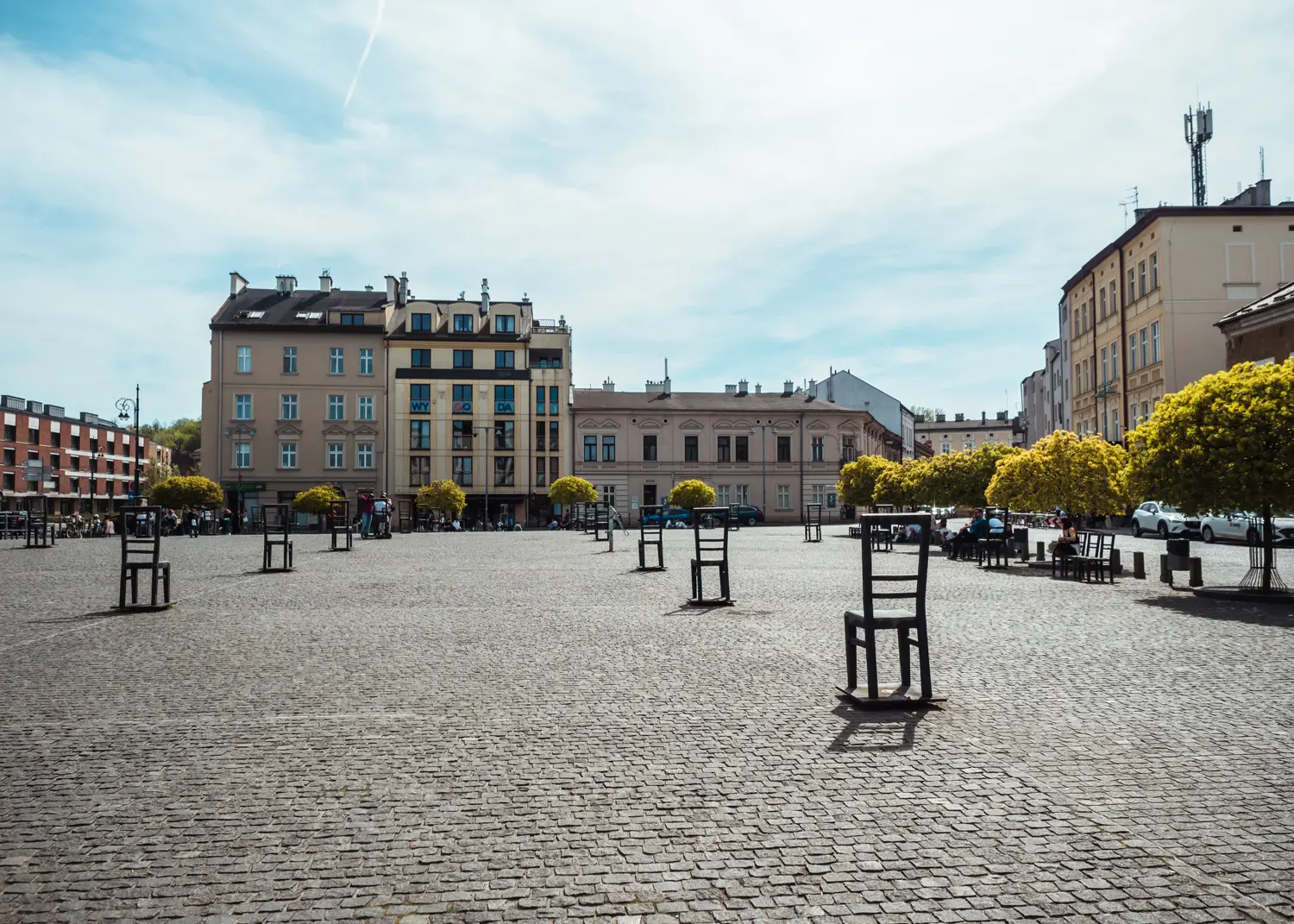 Chairs standing in line on the large, cobbled Ghetto Heroes square, surrounded by round trees and old apartment buildings in Podgorze, one of the most affordable areas to stay in Krakow.