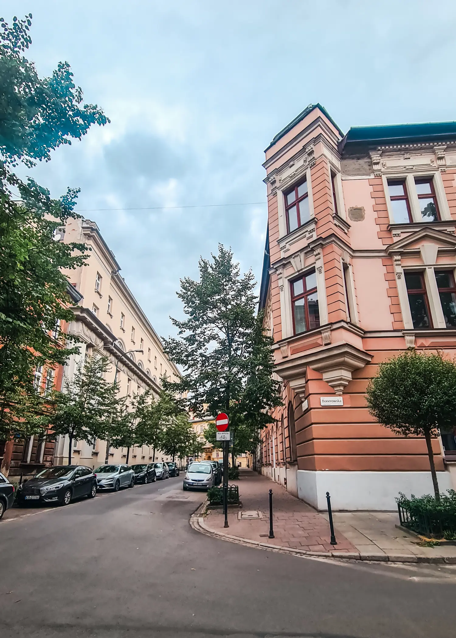 Streets lined with cars and trees, between two beige beautiful old buildings, on a cloudy day in Wesola, one of the best areas in Krakow.