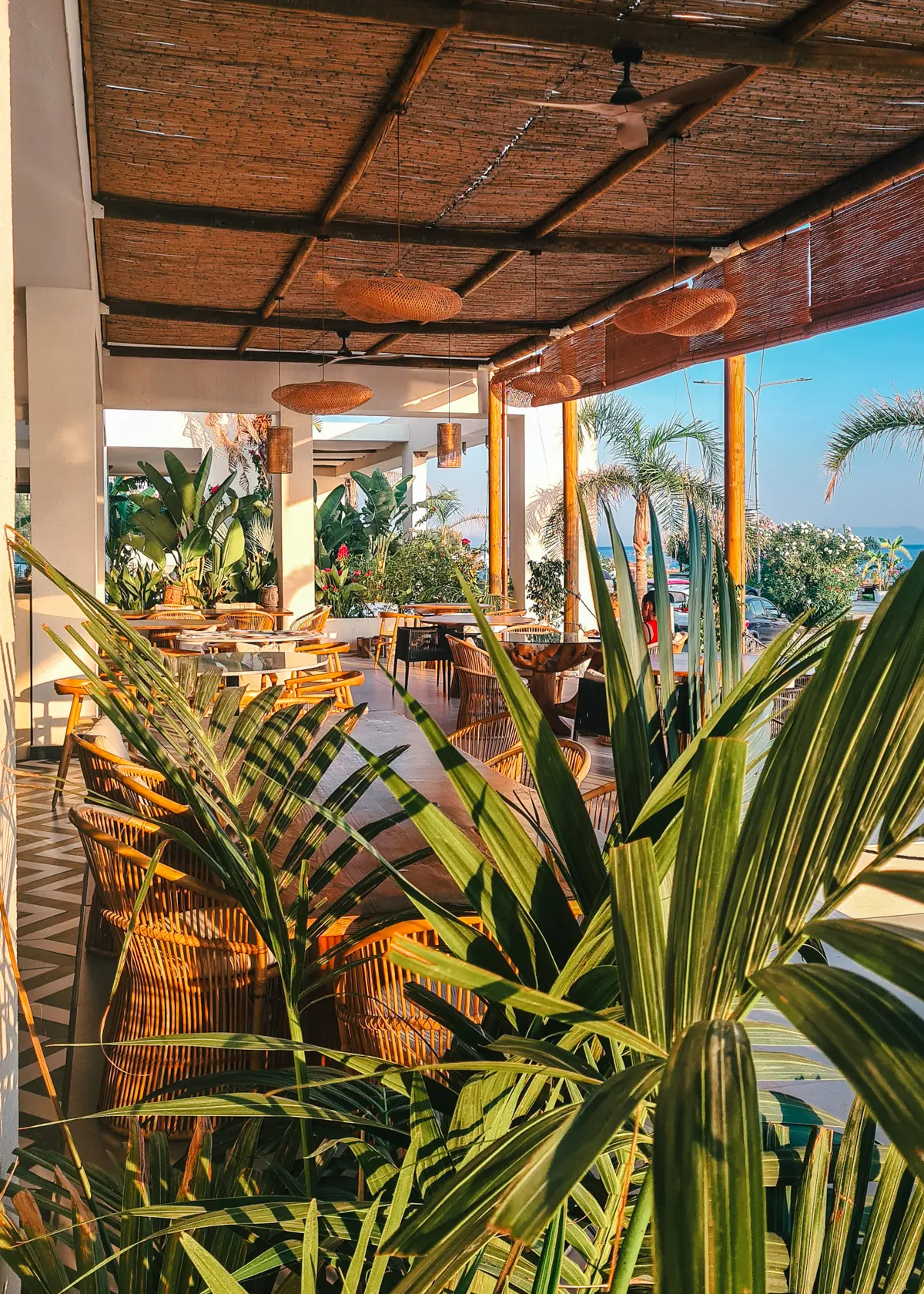 Looking over a small palm into a Bali-style restaurant with straw lamps, black and white floor and wooden tables at Abonora Restaurant at Drymades Beach in Albania.