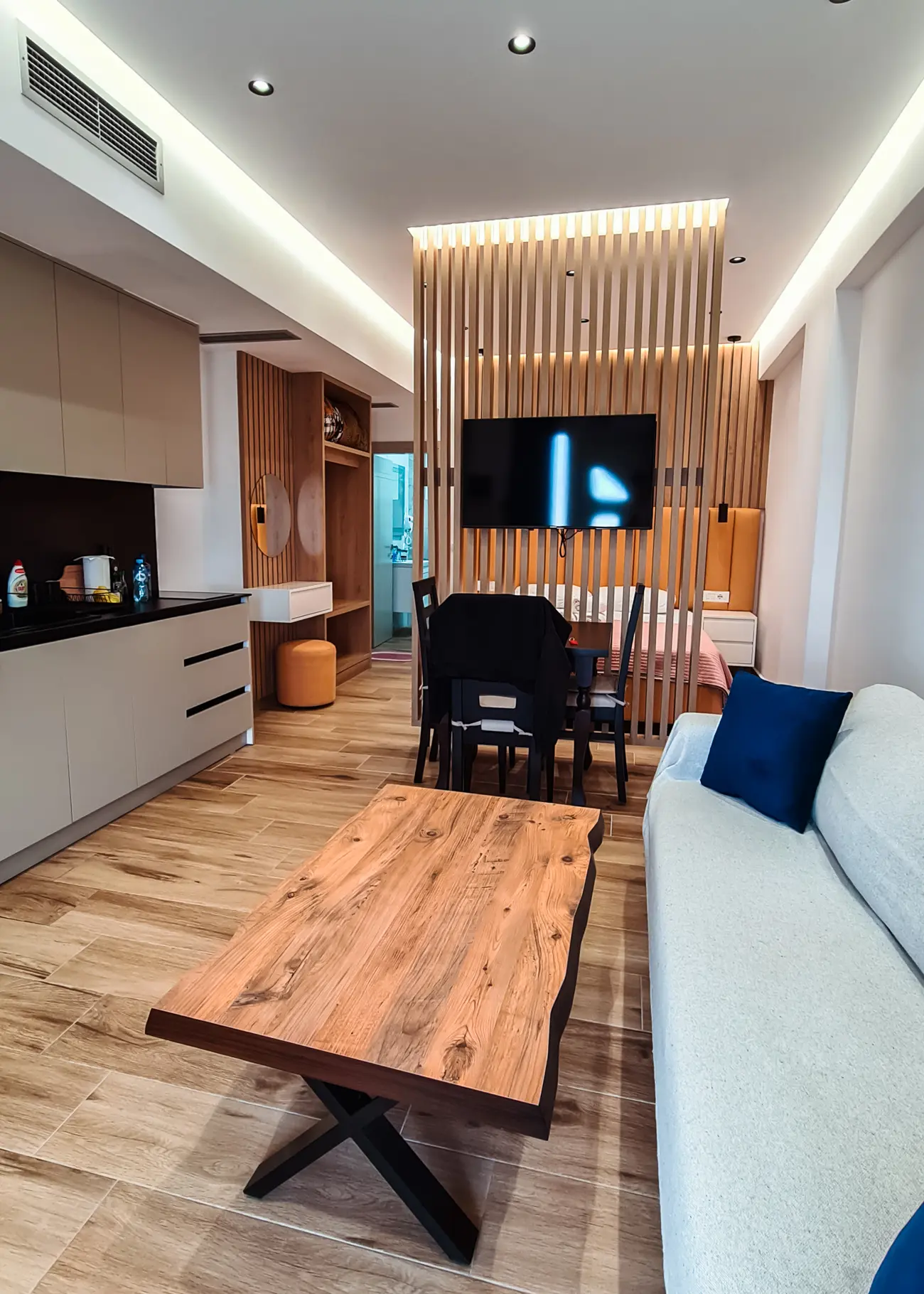 View of studio apartment with a beige kitchen on the left wall, wooden table in the middle and grey sofa on the right, with a wooden diving wall in the background in Sarand&euml;, Albania.