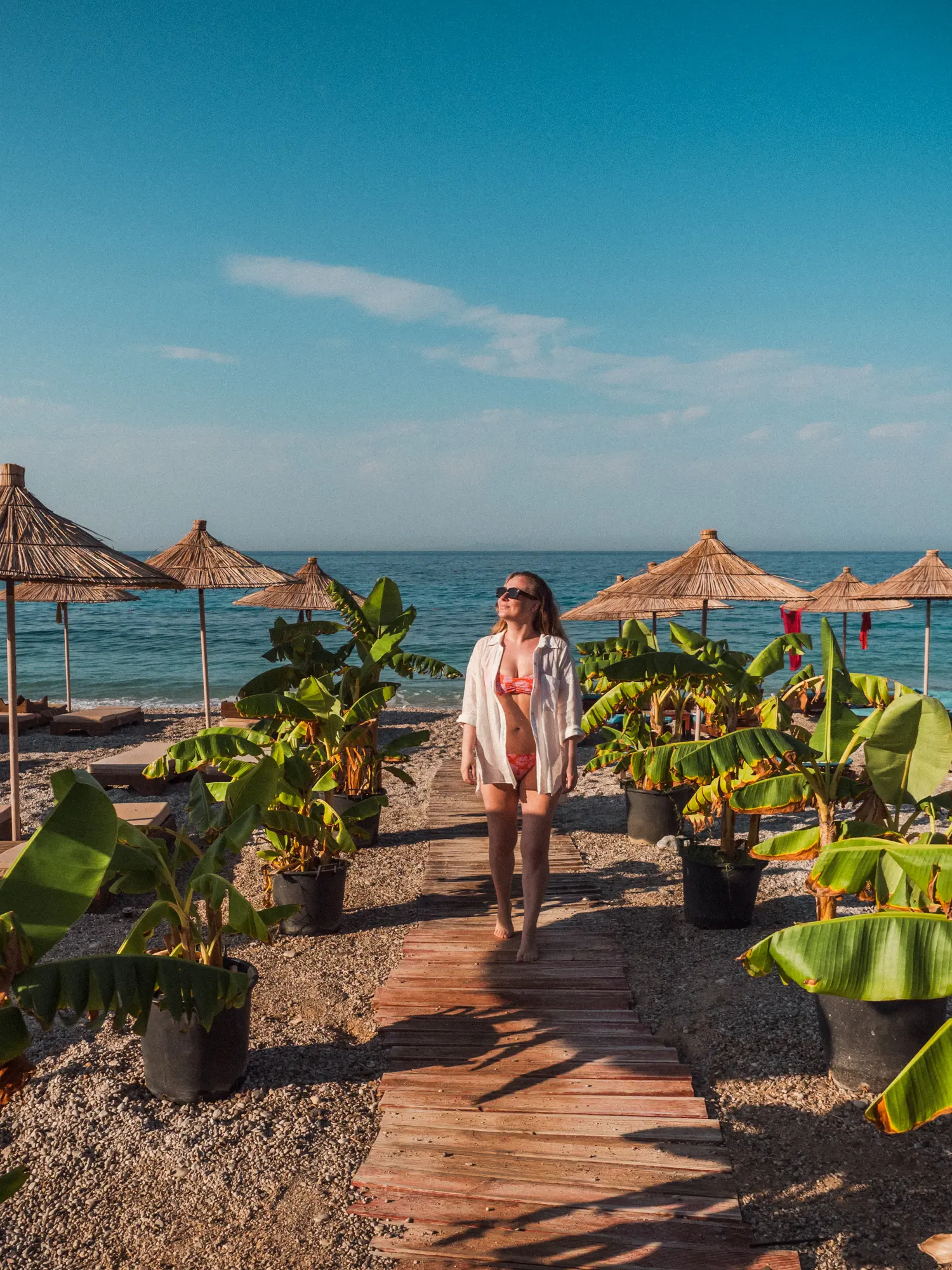 Woman wearing a pink bikini and open white shirt walking up a wooden path on the beach with banana palms and straw umbrellas on both sides at Drymades Beach in Albania.