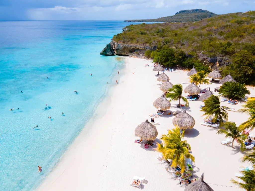 Drone view of the white-sand Cas Abao Beach with straw umbrellas and palm trees, people swimming int he light turquoise ocean and a green hill in the background, Curaçao.