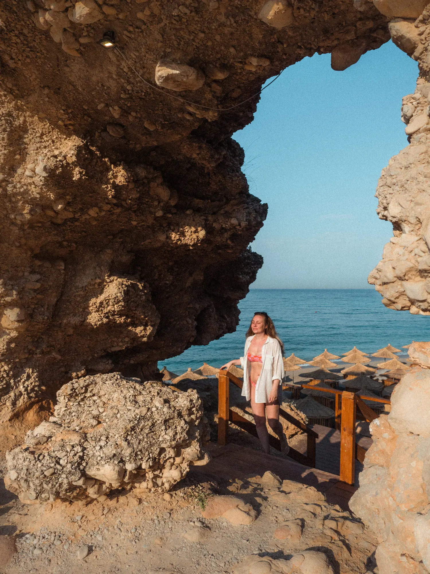 Woman wearing a pink bikini and open white shirt walking up the stair from a beach bar under a natural stone arch on Drymades Beach in Albania.