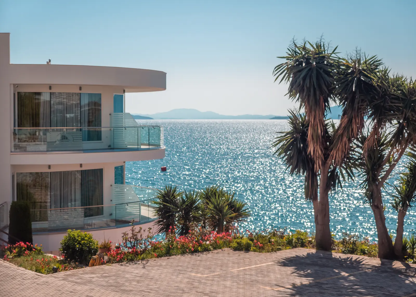 View from a street of the glistening ocean through the white curved Hotel Delfini in Saranda on the left and a palm tree on the right.