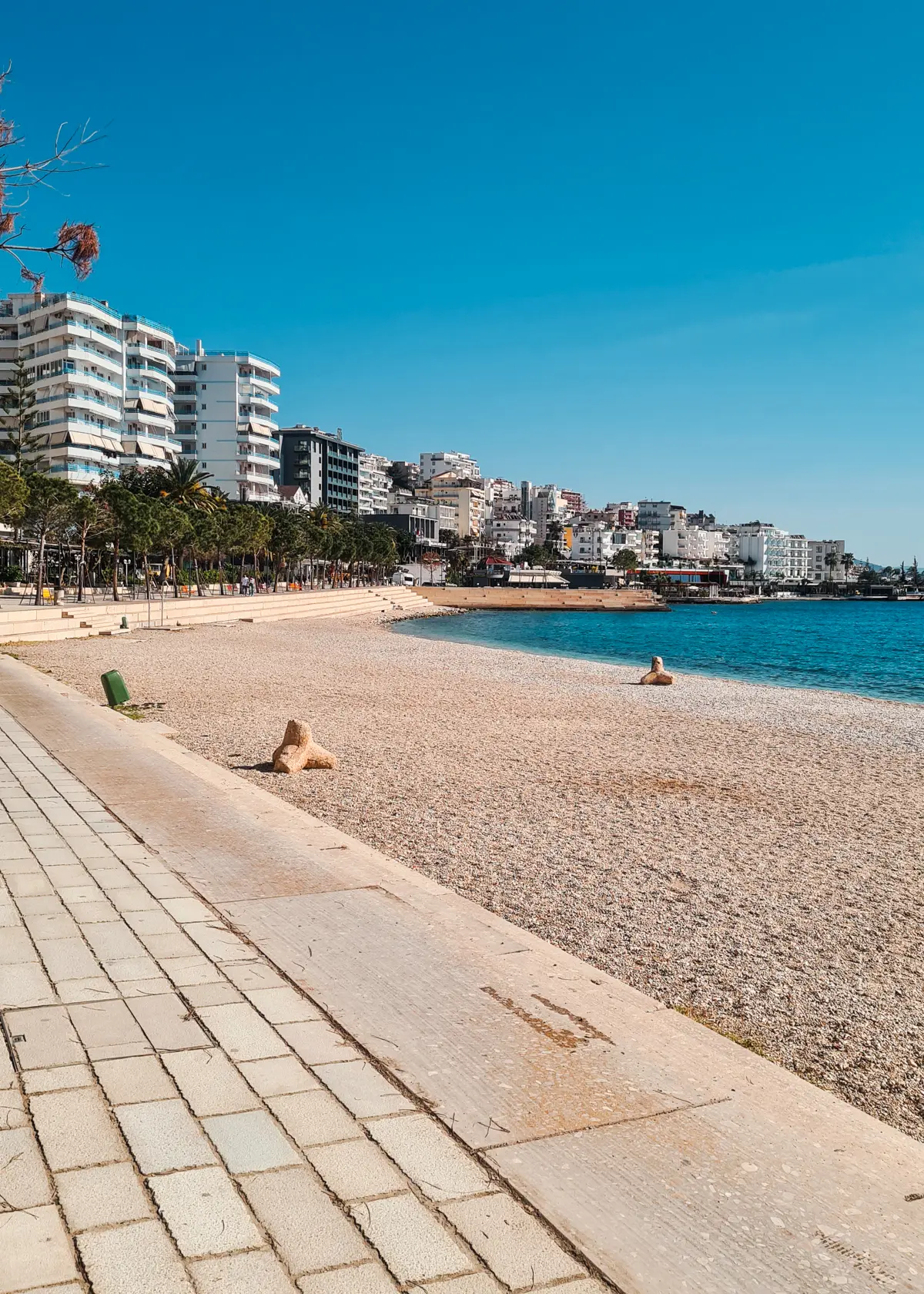 View of the pebble public beach in Saranda next to the promenade with a few high-rise buildings in the background.