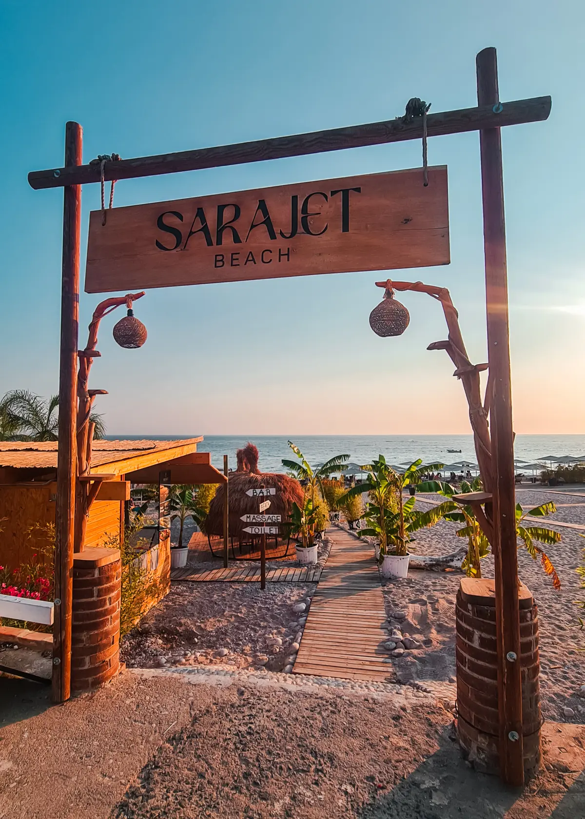 Wooden arch and sign at the Sarajet Beach Club with a wooden pathway and small palms on the beach in the background at sunset on Drymades Beach.